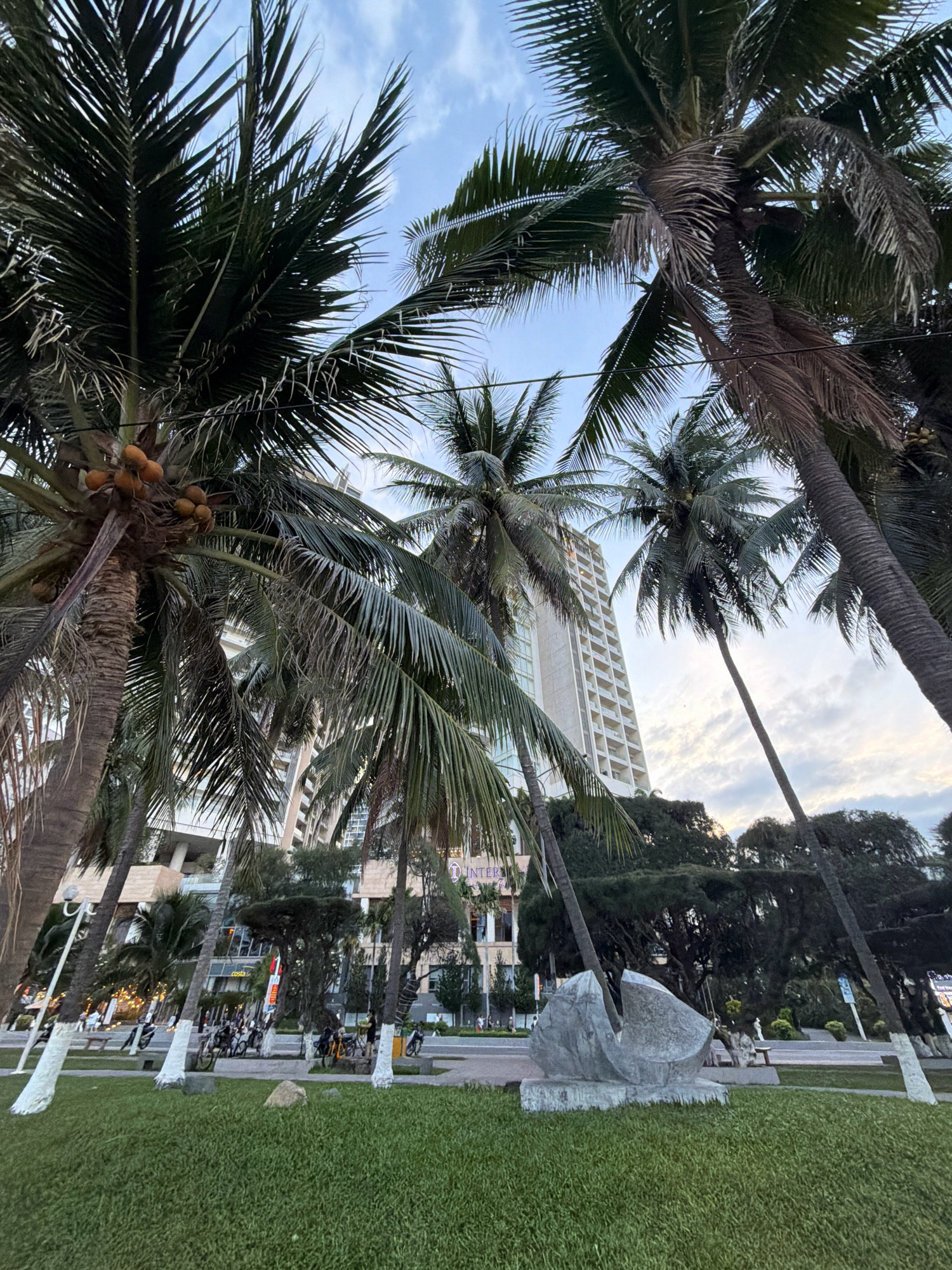 The image depicts a low-angle view of palm trees in the foreground, with a modern, tall building visible in the background. The building is light-colored with numerous balconies and windows, suggesting a hotel or apartment complex. A street with some traffic and pedestrians is present behind the palm trees, and a well-maintained grassy area with a sculpture is in the middle ground. The sky is overcast with some clouds. The presence of palm trees, a tall building, and a street with pedestrian activity are consistent with a coastal urban environment. Based on the visual cues, this scene is likely located in Nha Trang, Vietnam, a city known for its beaches, resorts, and modern architecture.