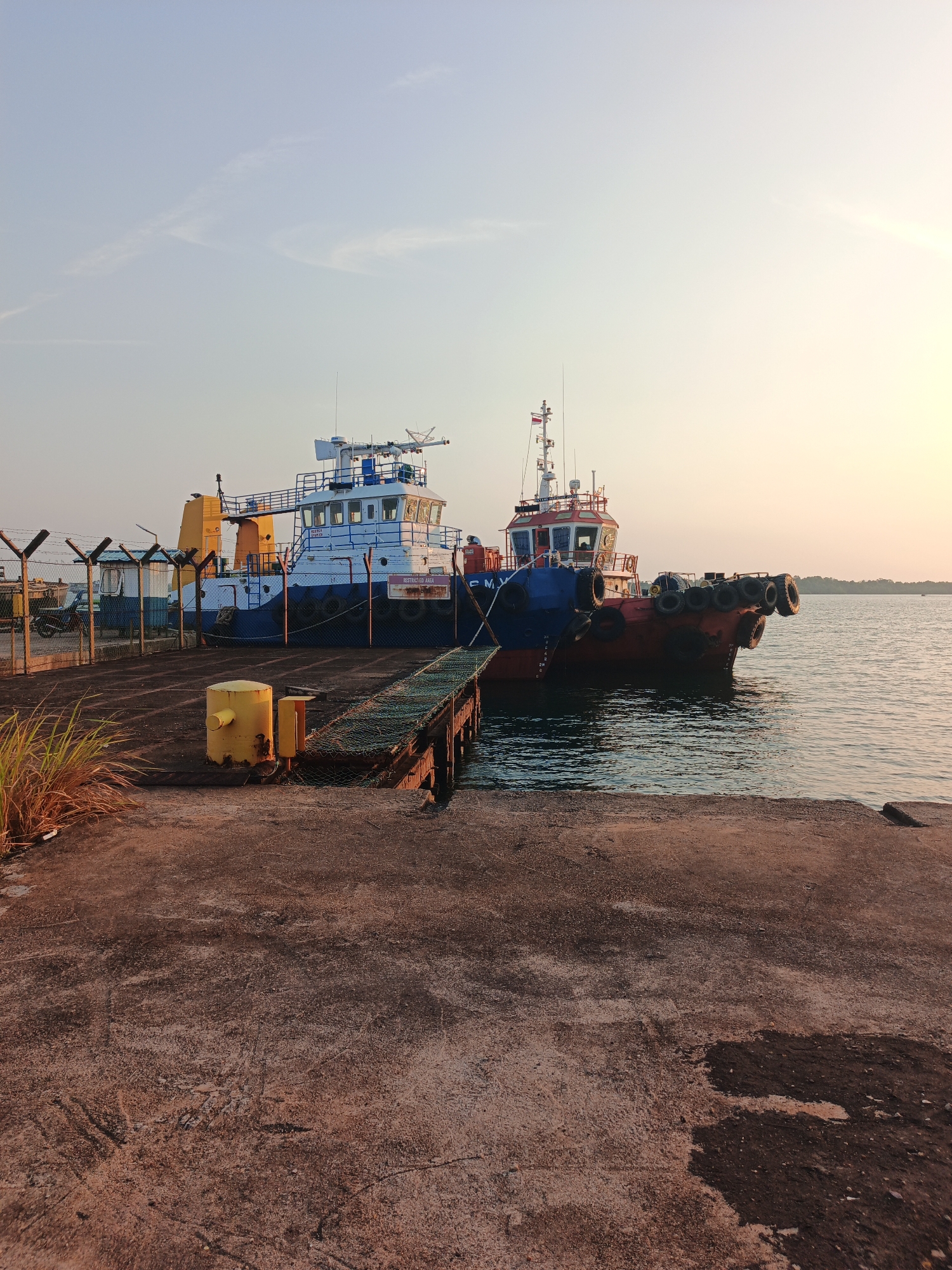This image depicts two tugboats docked at a pier in Batam, Indonesia, during a sunrise or sunset. The sky is a soft gradient of light blue and pale yellow, with wispy clouds. The tugboats are positioned side-by-side, with the one on the left being larger and painted blue and white, while the one on the right is smaller and predominantly red and blue. Both boats have visible radar equipment and masts.
The pier itself is made of concrete, with a textured and weathered surface. A metallic walkway, partially covered in netting, leads from the pier to the boats. In the foreground, a yellow bollard stands prominently on the concrete. To the left of the bollard, some dry, wispy grass is visible. Beyond the pier and to the left, a chain-link fence with barbed wire on top separates the dock from a small building and a parked motorcycle, suggesting a functional, possibly industrial, area. The water is calm, reflecting the warm light of the sky. The overall atmosphere is serene and quiet, indicative of an early morning or late evening at a port.