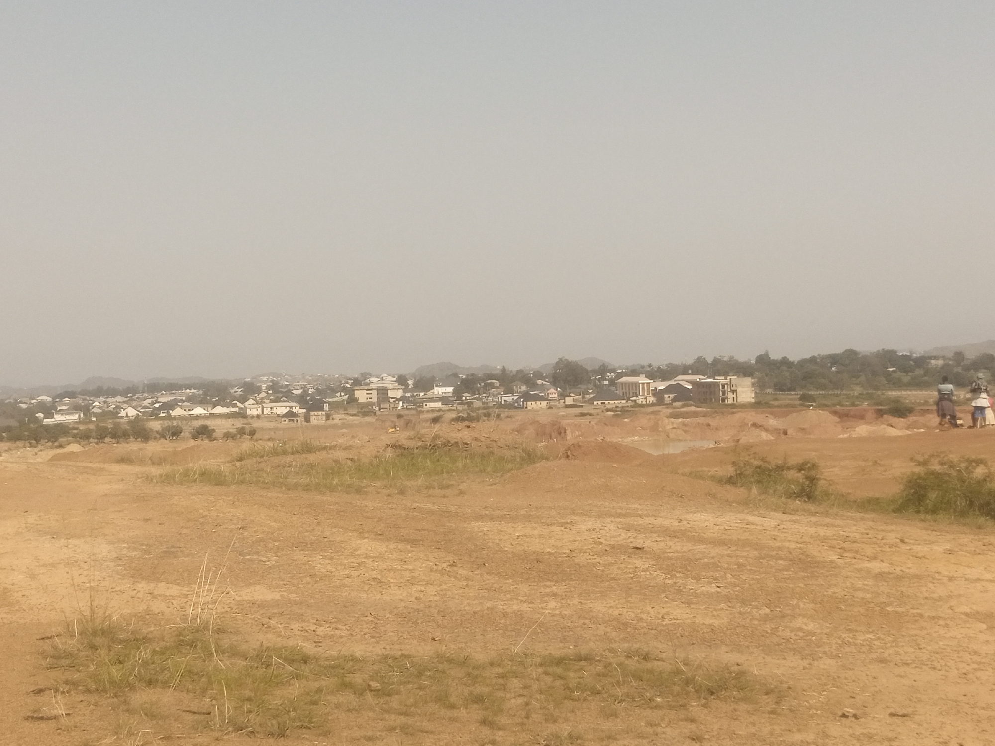 The image captures an overview of a developing area in Jos, Nigeria. In the foreground, a dry, dusty terrain with sparse dry grass dominates the landscape. The midground reveals a construction site with mounds of red earth, suggesting excavation or earthmoving activities. Several buildings, varying in architectural style and size, are visible in the background, indicating a growing urban settlement. Some of these buildings appear to be modern residential or commercial structures. A few individuals, two women and one man, are present in the midground, standing near the construction site. The atmosphere appears hazy, possibly due to dust or pollution, which is common in the dry season in Jos. The overall scene depicts a transition zone between undeveloped land and urban expansion.