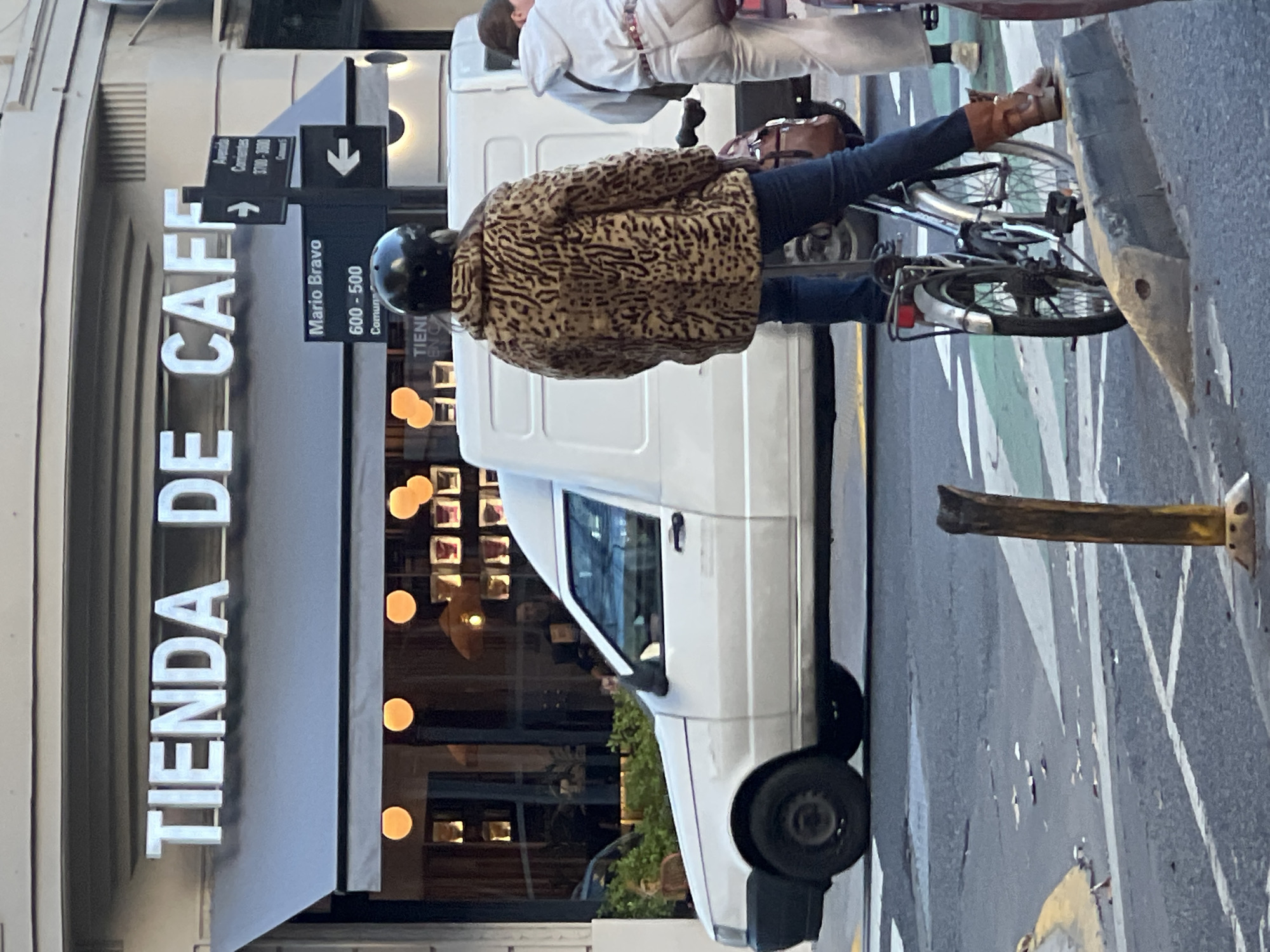 A daytime street scene shows a person wearing a leopard-print jacket, dark pants, and brown shoes, riding a bicycle on the right side of the frame. The person also wears a dark helmet. They are riding alongside a white van. To the left, a storefront is visible with the large text "TIENDA DE CAFE." A sign on the left reads "Mario Bravo 600-500, Corrientes 3700-3600." These are street names and number ranges in Buenos Aires, Argentina. The storefront is likely a cafe. A second person is slightly visible riding in the background. A yellow sidewalk marker is in the bottom-right foreground. The street and storefront are in focus, while the sky is blurred.