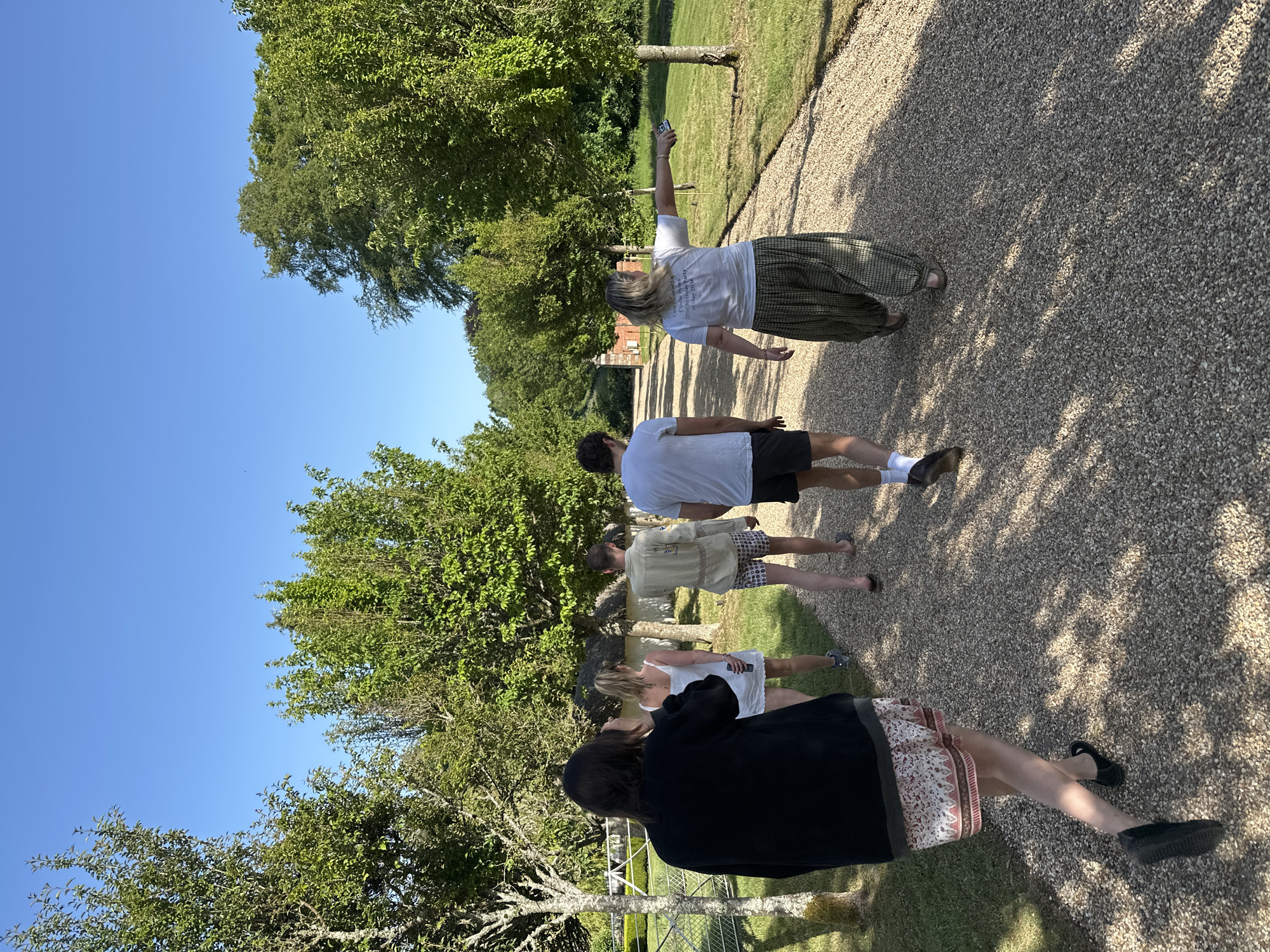 The image shows a group of five people walking along a gravel path in an outdoor setting. The sky is clear and blue, with trees lining both sides of the path.
From the back, the people include:
1. A person in a white shirt and patterned skirt is holding a phone above their head, potentially taking a picture.
2. A person wearing a white shirt and dark shorts walks further down the path.
3. A person in a light-colored jacket and patterned shorts is walking closely behind.
4. A person wearing white.
5. A person in a black shirt and patterned skirt walks in front of the group.
The environment includes trees, a clear blue sky, and a path covered in small gravel. The location is potentially a rural area, given the greenery and type of path.