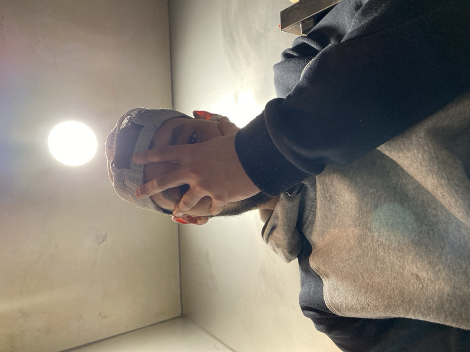 A person wearing a gray baseball cap and a gray and black hoodie covers part of their face with their hand. Their fingernails are painted red. A bright light is in the upper left corner of the image. The person is positioned in the corner of a room, with a gray wall visible in the background. The perspective is from below, looking up at the person.