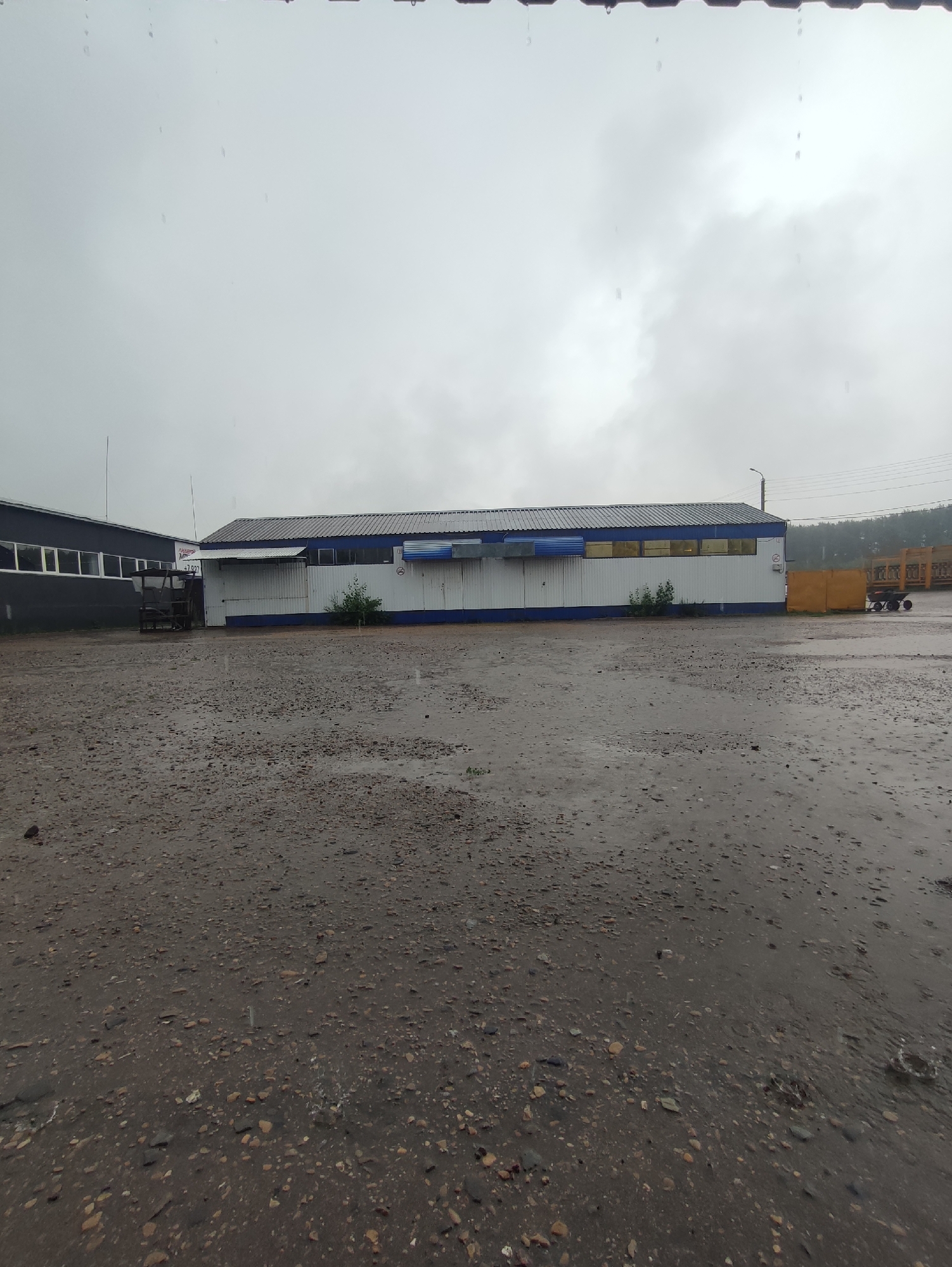This is a medium shot, exterior, eye-level, daylight photograph taken in Bakaly, Russia. The scene depicts a wet, gravelly outdoor area in front of an industrial building complex on a cloudy, rainy day. The ground is covered in small, varied-colored stones and is wet, with visible puddles and ripples from falling raindrops.
To the left, a dark gray industrial building with a row of windows is visible. Adjacent to it, a white industrial building with a blue trim and a corrugated metal extension is the main focus. This building has several windows with yellow or orange tinted glass, and a blue awning. There are a few sparse green bushes planted in front of this building.
Further to the right, a tall, bright yellow rectangular object stands at the edge of the property. Behind it, a portion of a wooden structure, possibly scaffolding or lumber, is visible. In the far distance, trees can be seen against the overcast sky. The sky is a uniform, pale gray, indicating overcast conditions and rain. The overall impression is one of a working industrial area experiencing inclement weather.
