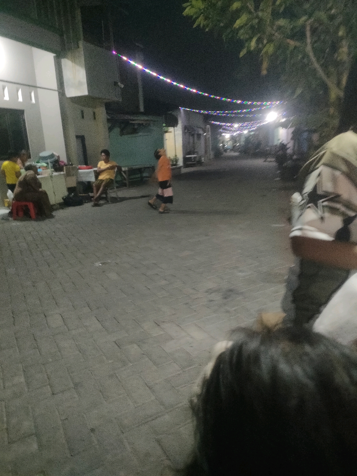 The image depicts a street scene at night in Surabaya, Indonesia, illuminated by strings of colorful lights strung across the street. In the foreground, a person's head and shoulders are visible from behind, appearing to be seated or looking down. The midground shows several individuals: a person in a yellow shirt, another sitting on a red stool, a person in an orange shirt walking, and another person sitting on a chair. Some objects that appear to be tables and chairs are also present. The background reveals buildings and trees, with more strings of lights extending into the distance. The street is paved with interlocking bricks. The overall impression is that of a community gathering or event taking place in a residential area.
