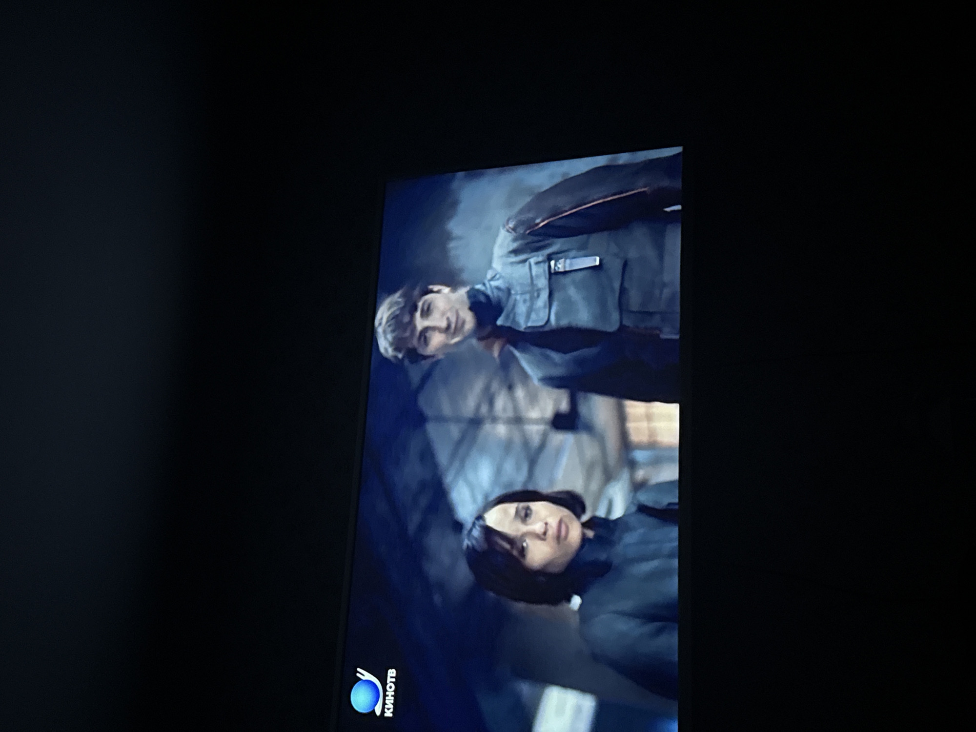 Here is a description of the image:
The image shows a television screen displaying two individuals. A man is seen in the upper part of the screen, wearing a gray uniform-like jacket with a badge on the chest. He is looking towards the left of the frame. Below him is a woman, also wearing a similar gray jacket. Her gaze is directed towards the viewer. The background is blurred and appears to be an urban or indoor setting. In the lower left corner of the screen, there is a logo that reads "Kиhотв." The television appears to be located in an indoor environment, based on the surrounding dark area. There is no information about Mytishchi, Russia, included in the image.