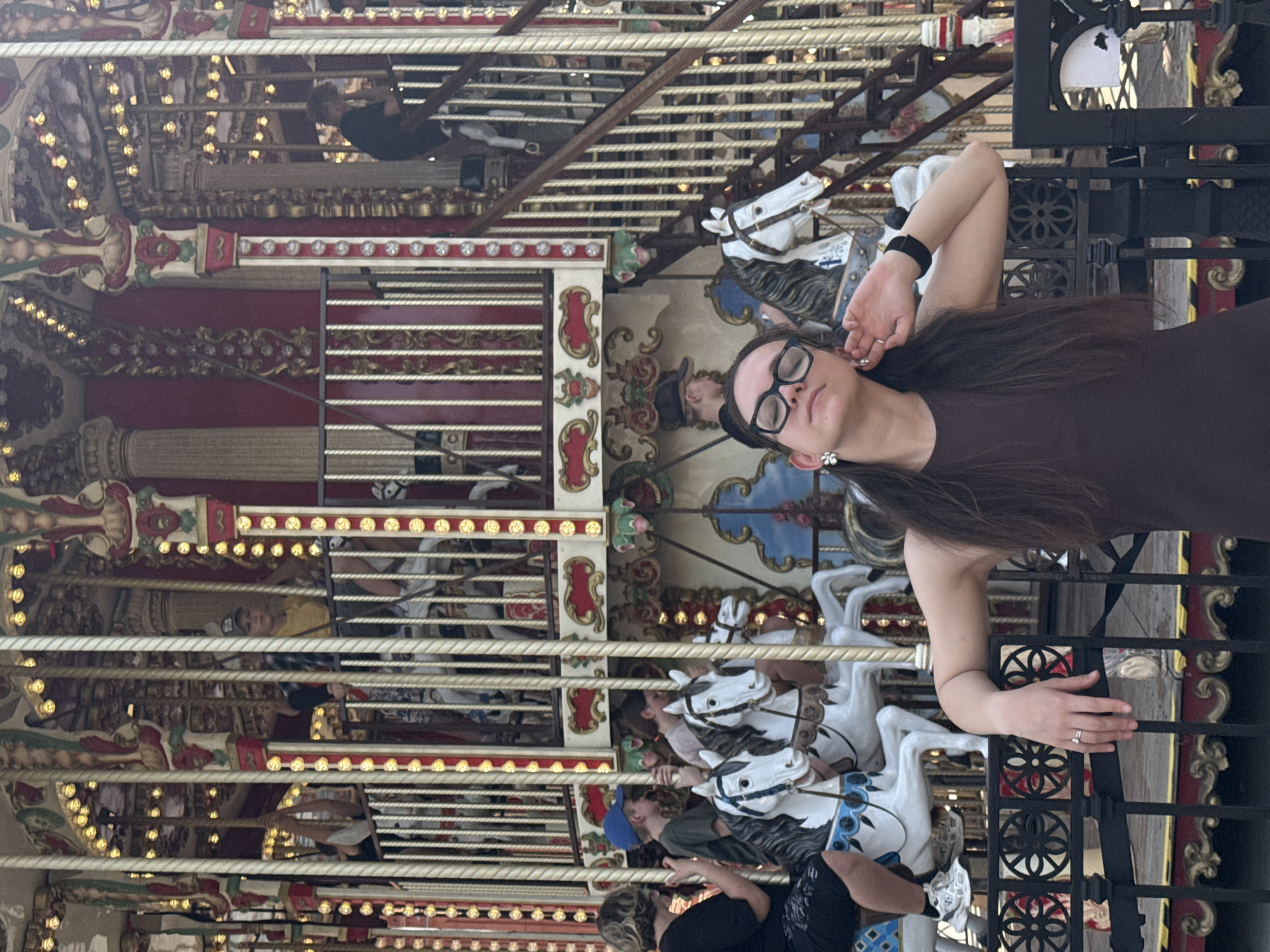 A woman with long brown hair and glasses leans against a black ornate metal fence in front of a carousel. The carousel features multiple rows of white horses adorned with blue and red detailing. Decorative elements include ornate red and gold trim, repeating patterns of lights, and painted scenes on the carousel's panels. People can be seen riding on the carousel. The location is Moscow, Russia.