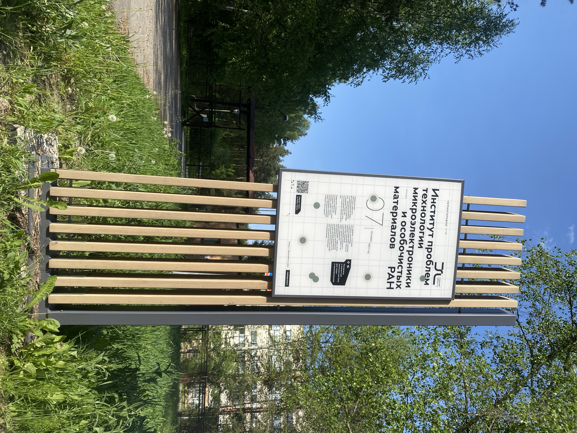 Here is a matter-of-fact description of the image:
The image shows a sign in a park-like setting, likely in the city of Chernogolovka, Unknown. The sign is mounted on a metal and wood frame. The sign itself has information written in Russian. The text includes "Институт проблем технологии микроэлектроники и особочистых материалов РАН" (Institute of Problems of Technology of Microelectronics and Especially Pure Materials of the Russian Academy of Sciences). Other text includes "Основан 1983 год" (Founded 1983). There's a QR code on the sign. The background consists of grass, trees, and a blue sky. A building is visible in the reflection of the sign. There's a path nearby.