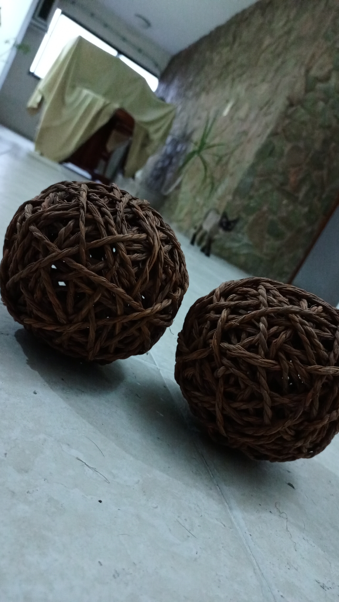 The image shows two brown, spherical objects made of interwoven material resting on a light-colored, tiled surface. The objects appear to be decorative balls. In the background, there is a covered object or piece of furniture and a wall with a textured, possibly stone-like surface. A window with natural light is also visible. There is no information in the image to indicate that the scene is located in the city of Los Robles, Venezuela. No people or actions are depicted.