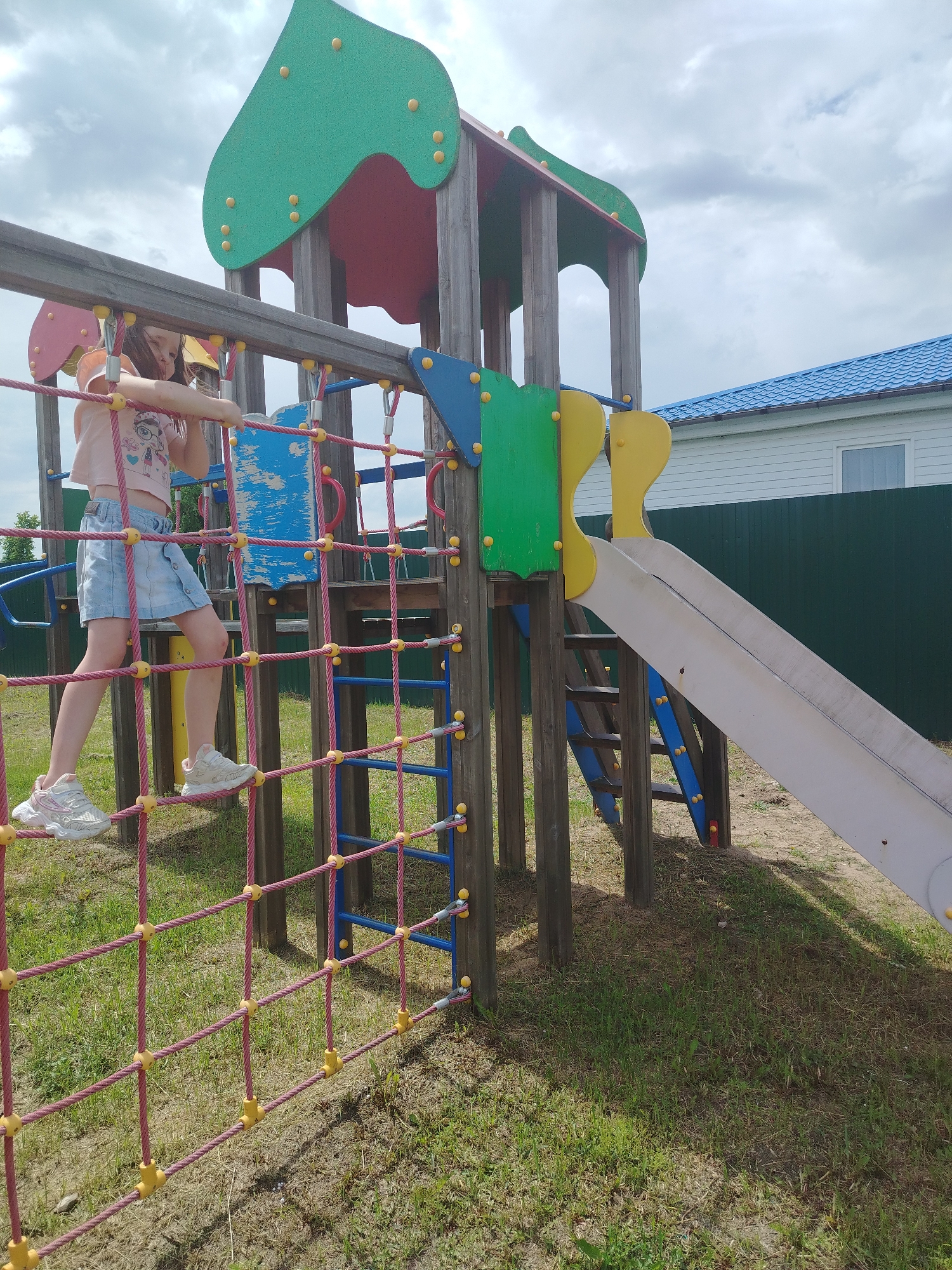 A young girl is on a playground climbing net made of pink rope and yellow connectors. She is wearing a light pink t-shirt and a light blue denim skirt, with white sneakers. The playground structure has wooden supports, colorful panels of green, yellow, and blue, and a white slide. Behind the playground is a green fence and a white building with a blue roof. The ground is covered in grass and sand. The scene appears to be a residential area in Klyuchi, Russia.