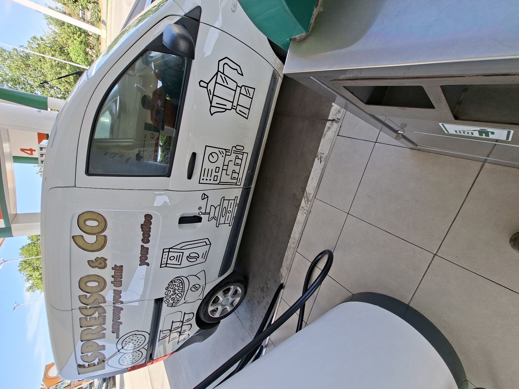 A white van with coffee-related decals and "Espresso & Co...anyway you drink your coffee" written on its side is parked next to a trash can at a gas station. The photo was likely taken in Plovdiv, Bulgaria, as the van appears to be at a gas station in a European city. There are no people visible outside the van. The immediate environment consists of the gas station pump and surrounding paved area.