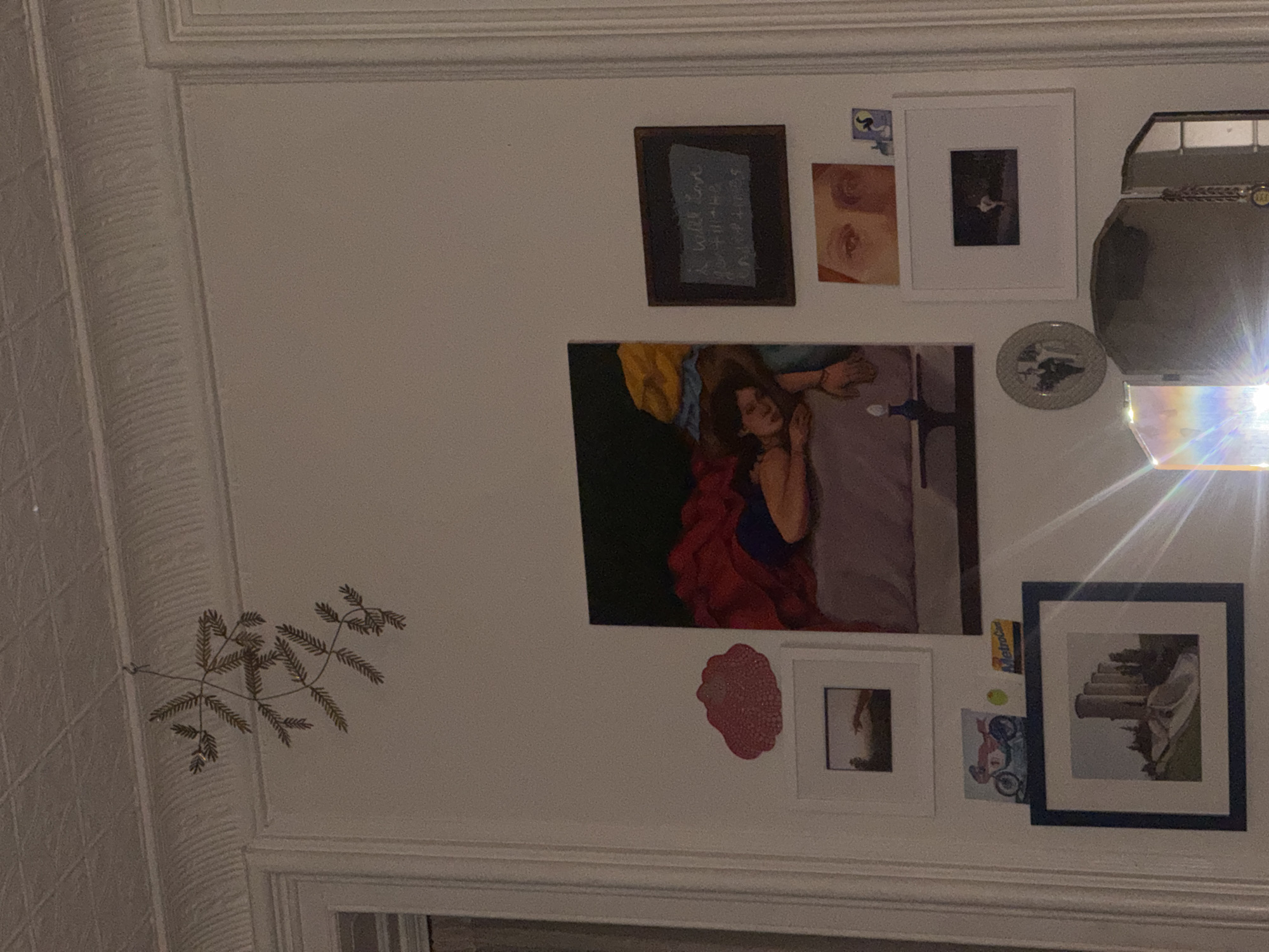 A section of a wall with a collection of artwork and decorative items is depicted. There are several framed pieces of art, including a portrait of a woman lying down, and other framed photographs and paintings. There is also a decorative metal branch attached to the wall. There is a ceiling and decorative molding present. The location appears to be an interior space. There are no people visible. The precise location in the city of New York is not discernible from the image.