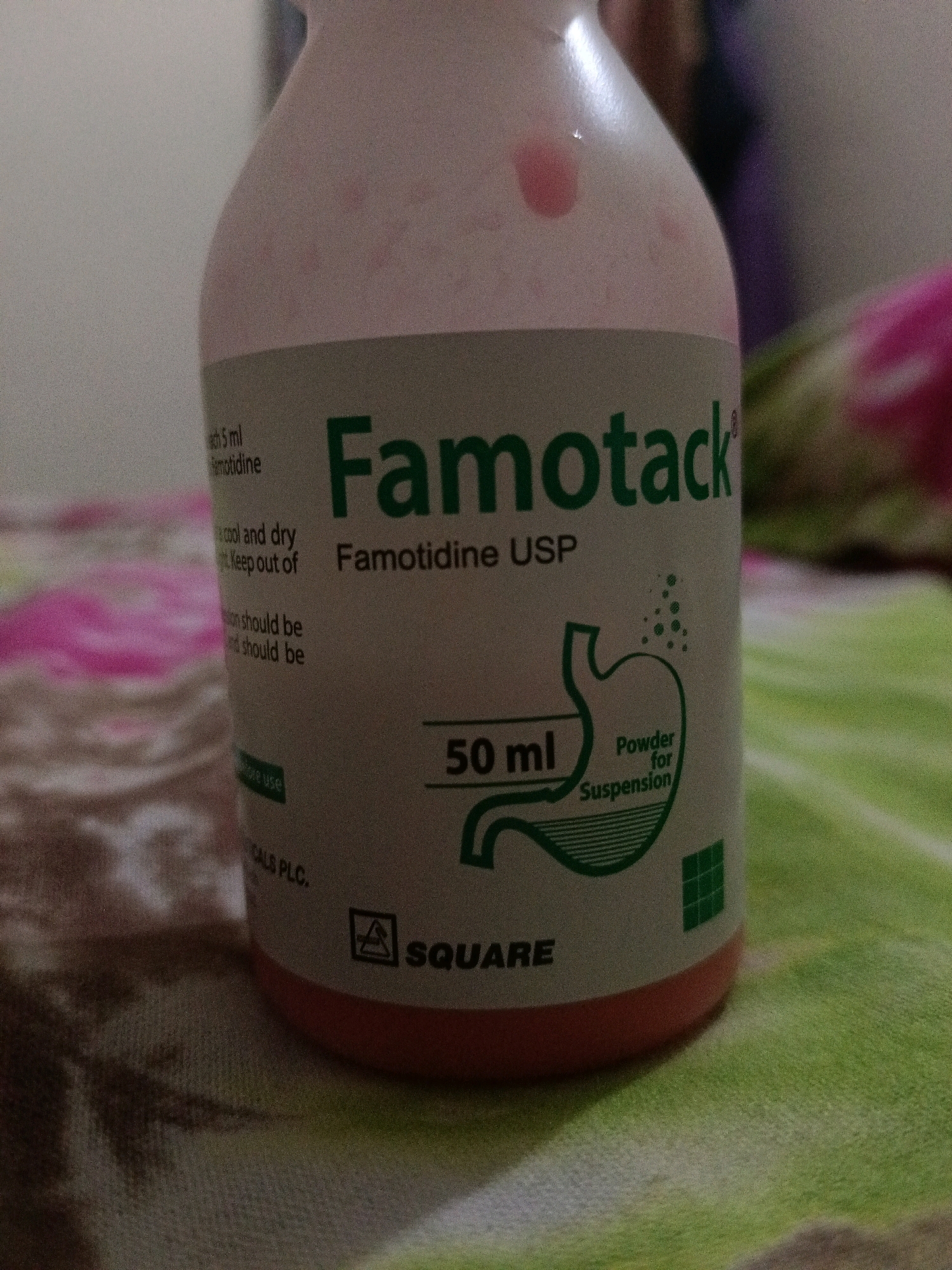 The image shows a close-up of a bottle of Famotidine USP powder for suspension. The bottle is light pink with a white label that has green text and graphics. The text on the label includes "Famotack", "Famotidine USP", "50 ml", "Powder for Suspension", and the Square Pharmaceuticals PLC logo. There are also some instructions written on the side of the label, which are partially visible and include phrases like "cool and dry" and "Keep out of". The bottle is placed on a patterned surface that appears to be a fabric, possibly a bedspread or a blanket. The background is blurred and out of focus, showing hints of other objects, including what appears to be clothing or fabric in shades of pink and purple. The lighting in the image is somewhat dim and diffused, creating a soft overall effect. The image does not provide any information about the city of Katiadi, Bangladesh, nor does it contain any discernible details about people or actions that would suggest a specific location or event.