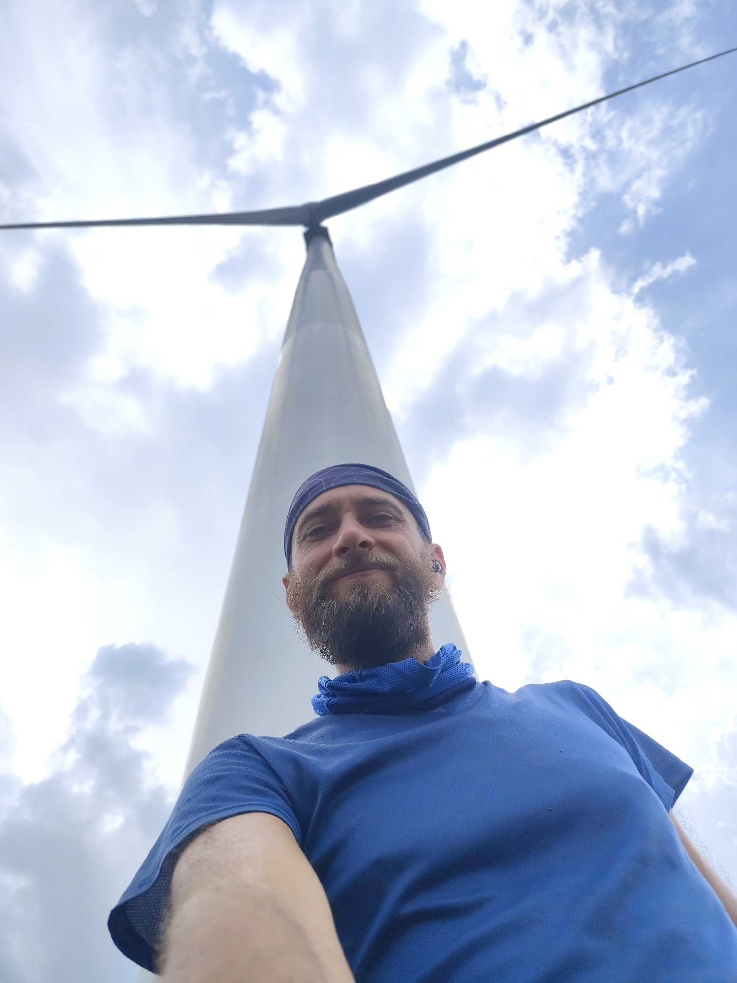 A man with a beard and a blue shirt stands in front of a wind turbine. He is wearing a blue head covering. The sky is cloudy. The photo appears to be taken from a low angle, looking up at the man and the turbine. The location is Phát Chi, Vietnam.