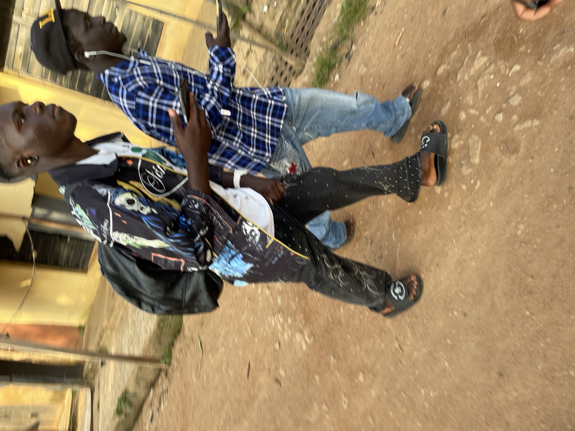 In Barakin Akawo, Nigeria, two young men walk on a dirt path. The man on the left wears a black backpack, a white t-shirt, and black pants with white dots and a graphic design. He has on black slides with a white emblem. The man on the right wears a blue and white plaid shirt, blue jeans with a rip on one knee, and black slides. Both men are looking forward and holding smartphones. The background shows buildings with yellow and orange walls, and some vegetation. The lighting suggests it is daytime.