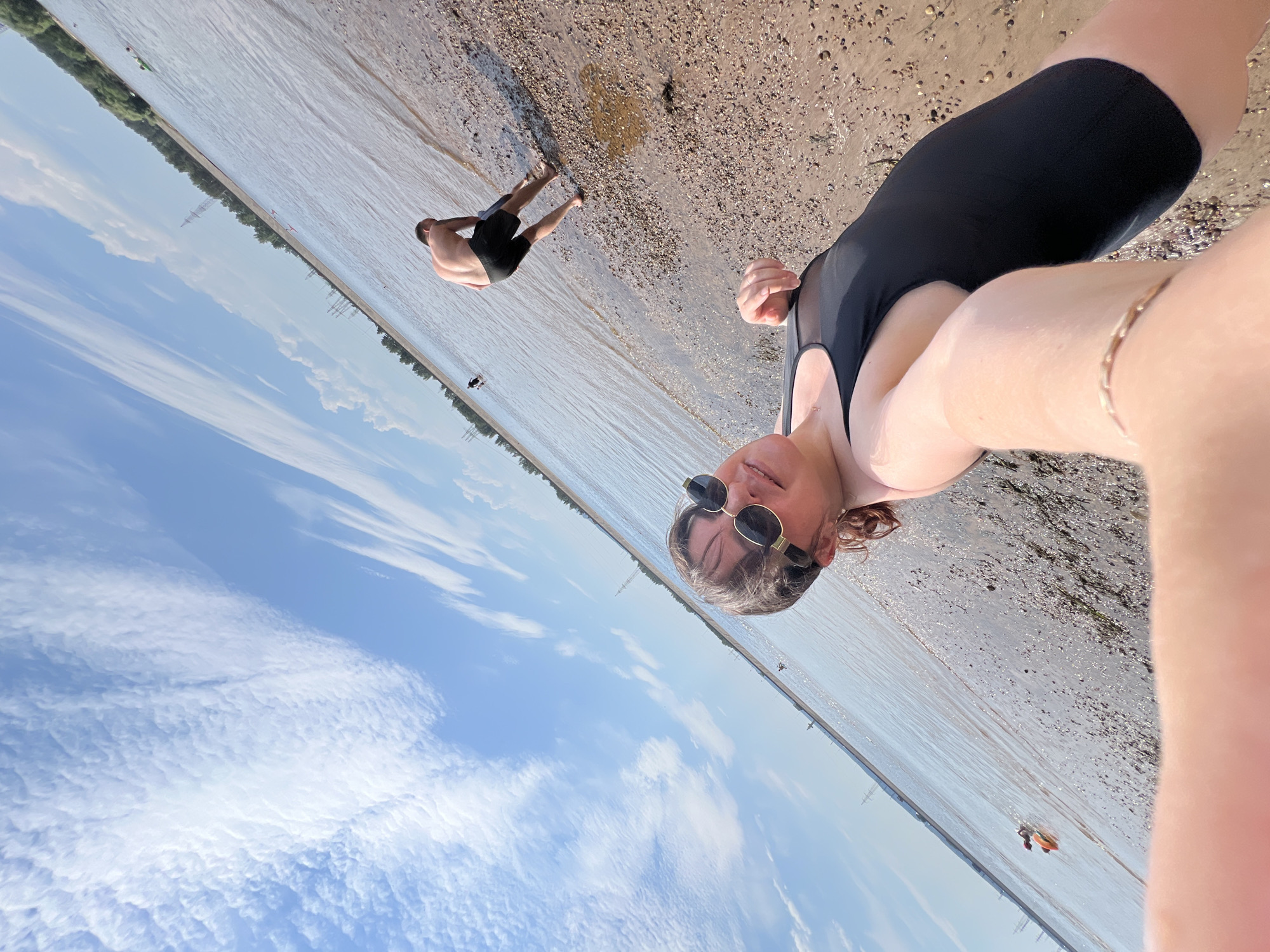 A selfie captures a woman in a black swimsuit and sunglasses at a beach with a body of water. She is in the foreground, and the camera's arm is visible. In the background, a man in black shorts stands near the water's edge, facing away from the camera, with his back to the viewer. The beach is composed of small stones and sand. The sky is blue with scattered white clouds. The location is identified as Noviy, Russia, suggesting a waterfront area within the city. The overall scene depicts a leisurely outdoor activity by the water on a sunny day.