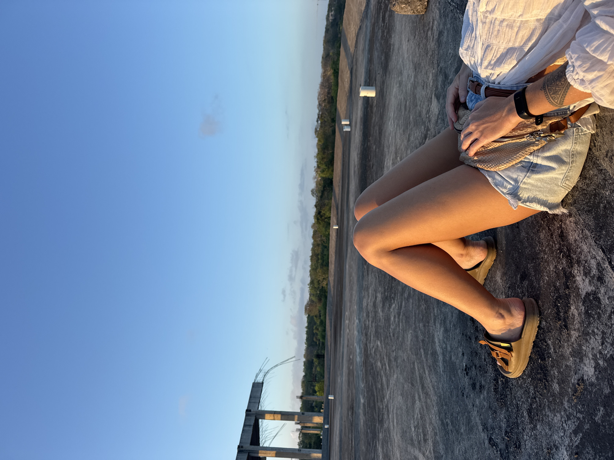 A person is lying down on a concrete surface, viewed from a high angle. They are wearing denim shorts, a white shirt, and brown sandals. A tan fanny pack is slung across their lap, and a black watch is visible on their left wrist. Their right hand rests on the fanny pack. In the background, there is a partially constructed concrete building and a line of trees under a clear blue sky with a few scattered clouds. The location is identified as Kuta Selatan, Indonesia.