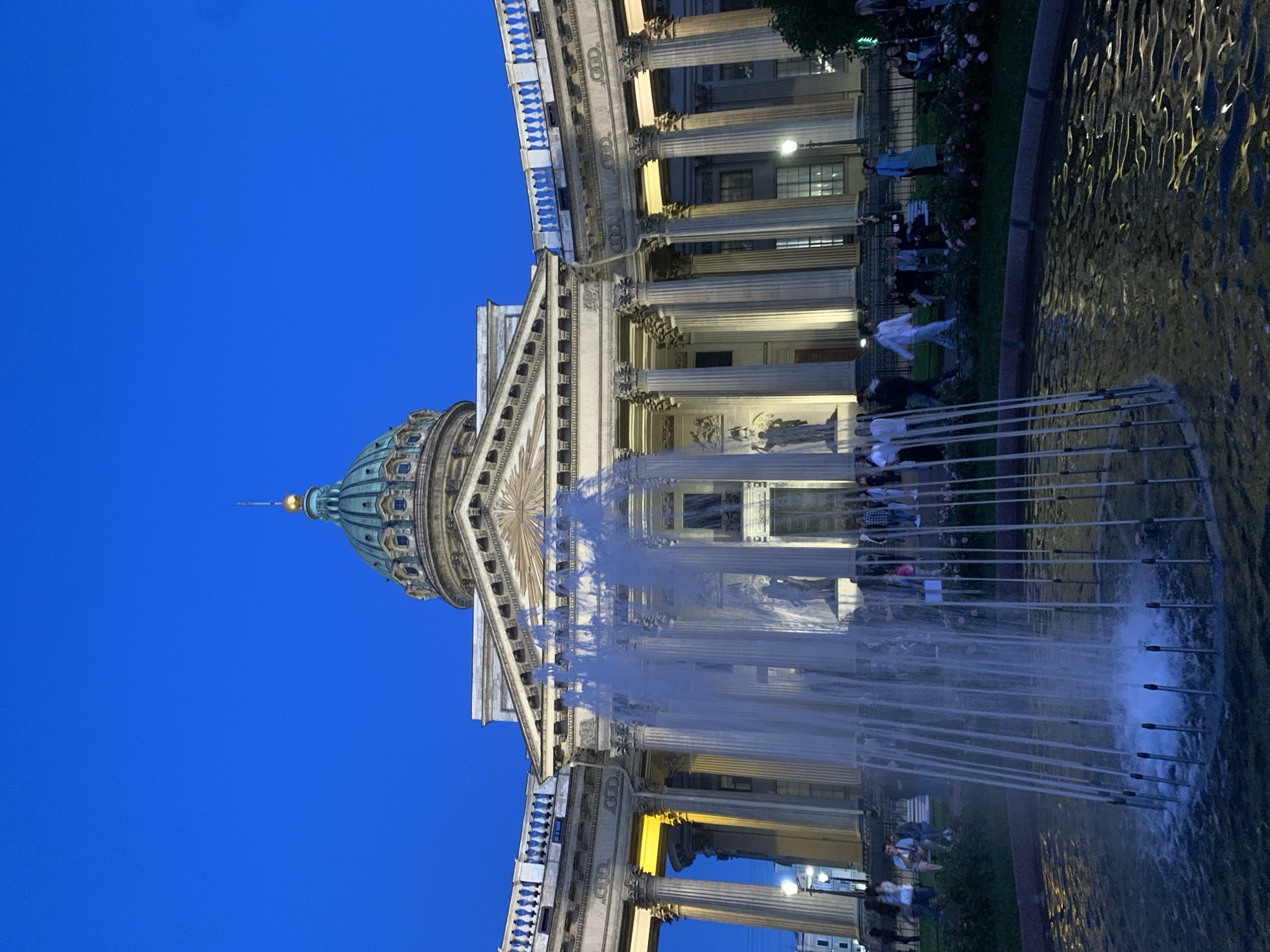 The image shows Kazan Cathedral in Saint Petersburg, Russia, at dusk. A fountain is in the foreground with water spraying upwards. Behind the fountain, the cathedral's grand facade is visible, featuring a dome, colonnades, and statues. People are gathered on the steps and in front of the cathedral, some sitting on benches and others walking. The sky is a deep blue, indicating twilight. The lighting suggests the presence of artificial lights illuminating the architecture. The atmosphere appears calm and public, with individuals enjoying the exterior of the landmark.