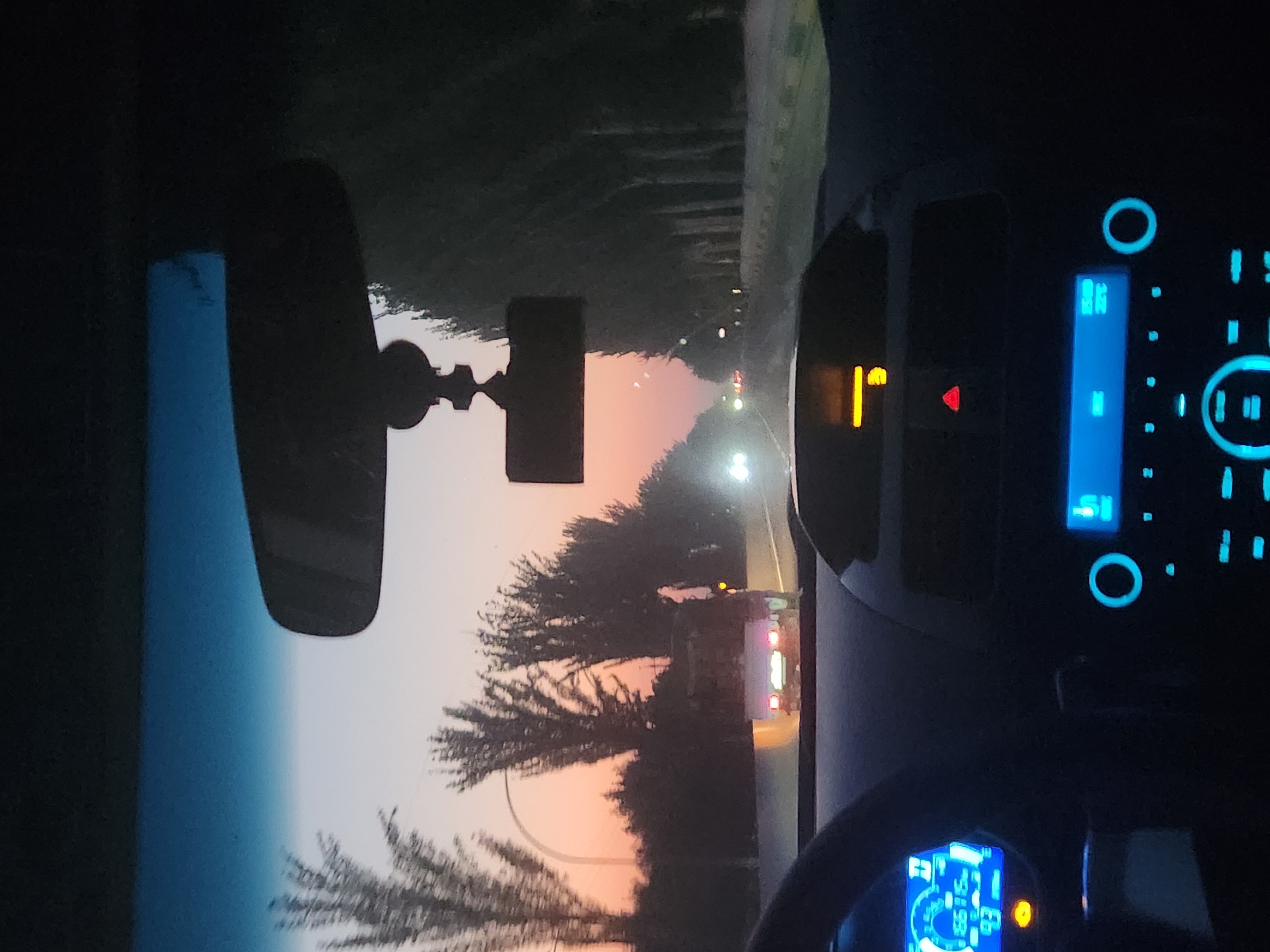 The image depicts the interior of a car driving on a road at dusk, with trees lining the right side of the road and a soft orange and blue gradient in the sky. The view is from the driver's perspective, showing the dashboard with illuminated displays. On the left side of the dashboard, a digital speedometer shows a speed of 93 and another number, 66716. The center console features a radio display with illuminated blue buttons and a circular control knob. A small red triangle warning light is visible on the dashboard. In the distance, headlights of other vehicles can be seen on the road, and a truck with illuminated taillights is ahead. The rearview mirror hangs from the windshield, and a dashcam is mounted below it. The location is identified as Sariqmirzachek, Uzbekistan.