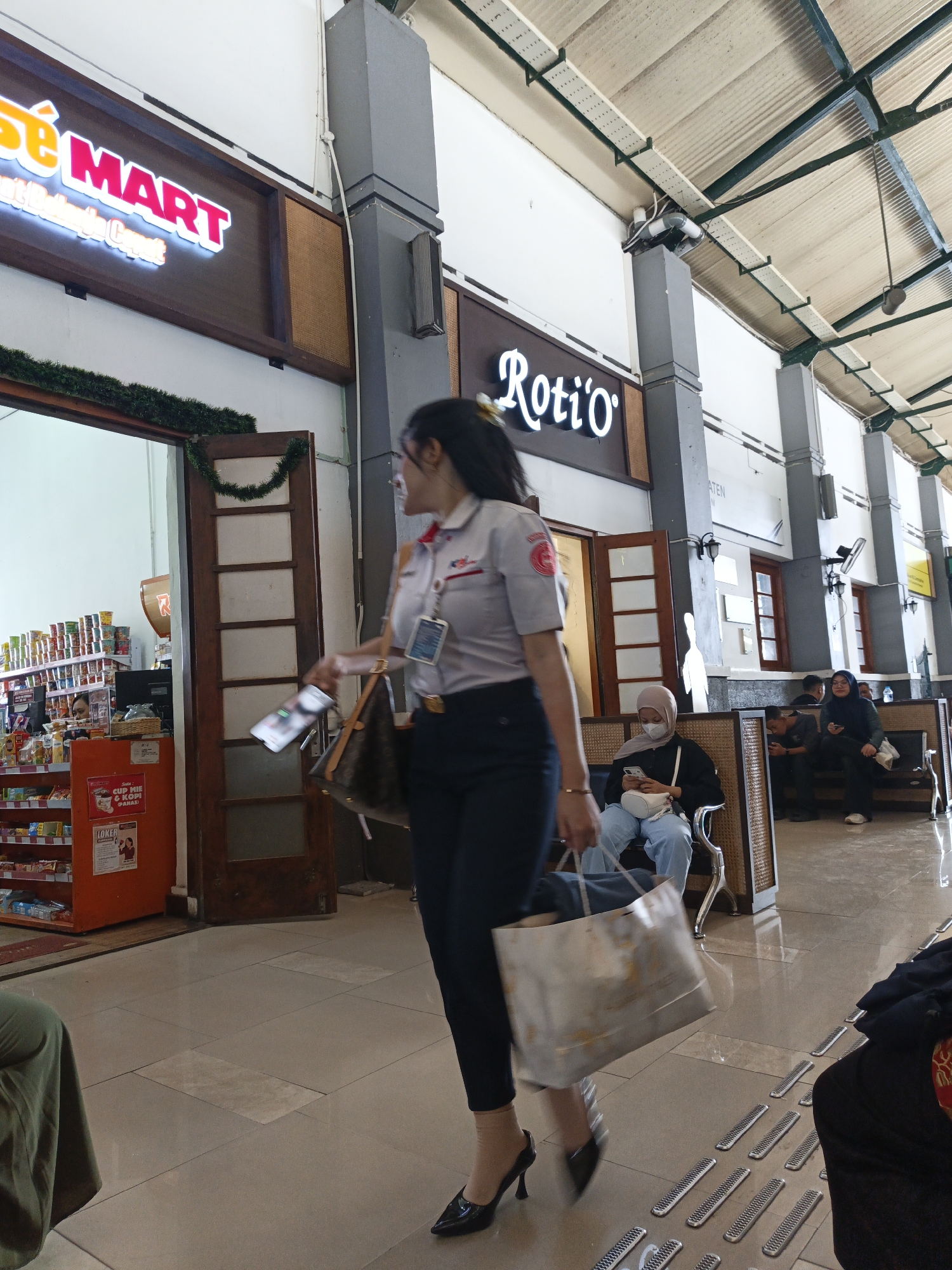 The image captures a scene at a train station in Klaten, Indonesia, likely within the waiting area or concourse. In the foreground, a woman in a grey uniform with a red and white logo is walking to the right, holding a paper shopping bag and a mobile phone. She is wearing dark trousers and black high-heeled shoes. To her left, a storefront sign reads "SE MART" in glowing neon letters. Further into the background, another neon sign displays "Roti'o". Several people are seated on benches in the waiting area, some of whom are looking at their phones. The environment appears to be an indoor public space with a tiled floor, pillars, and a covered ceiling with visible pipes and wiring. The overall impression is one of a busy transportation hub.