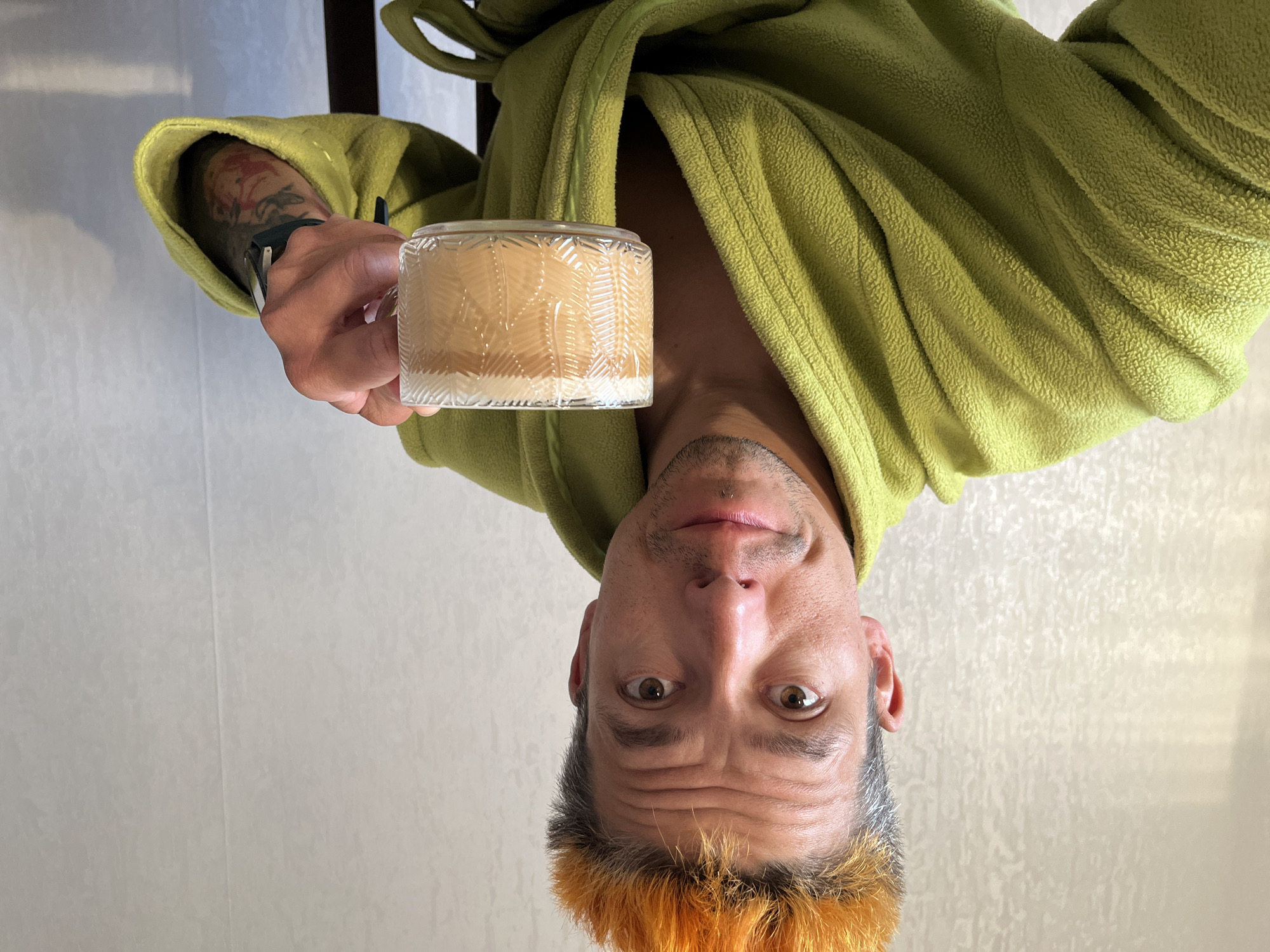 A person with short, two-toned orange and gray hair is holding a lit candle in a glass container. The person is wearing a green robe, a watch, and has tattoos on their left forearm. The background is a light-colored wall. The image is oriented upside down. There is no other information regarding location in Russia.