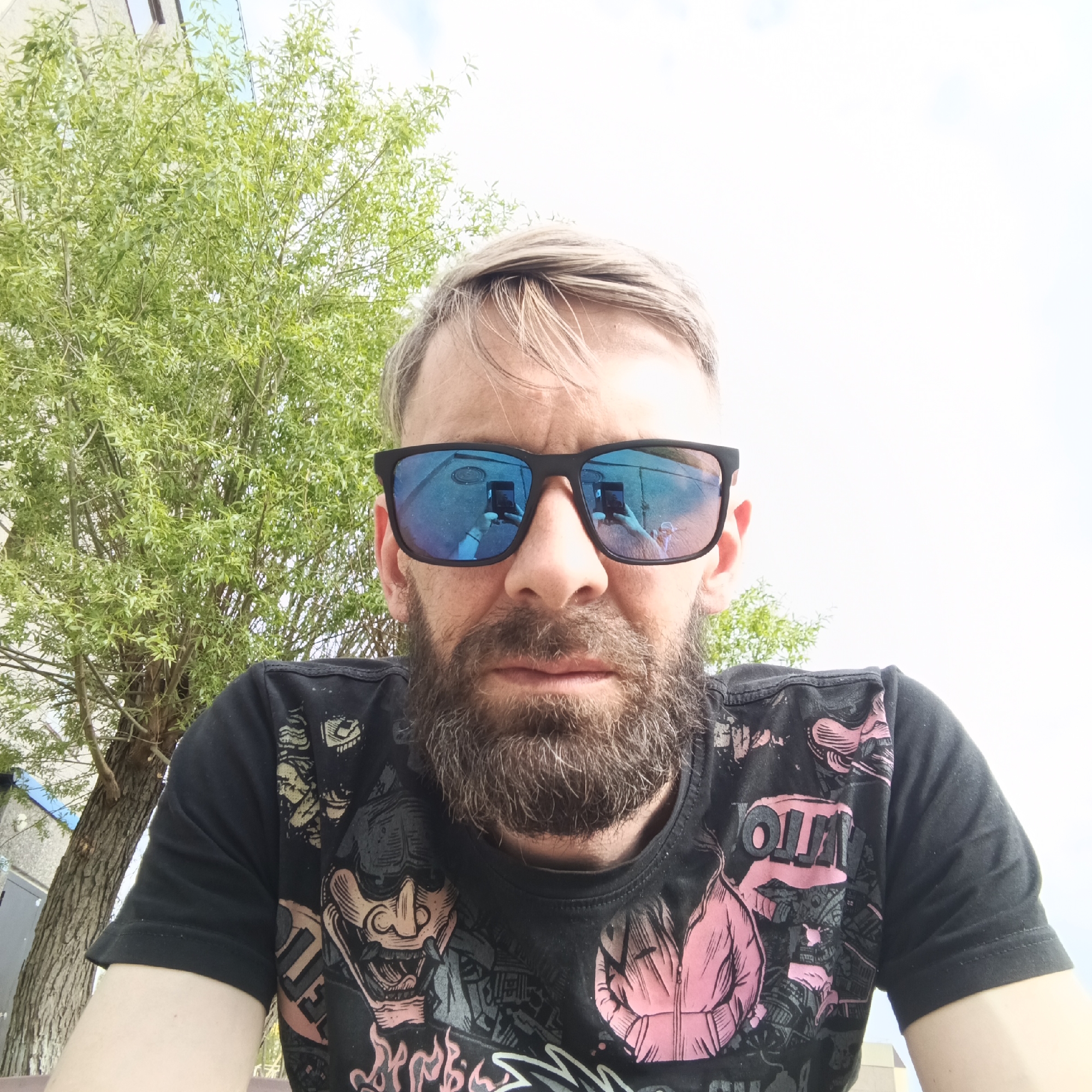 A man is centered in the frame, wearing blue reflective sunglasses that show a reflection of a cell phone being held in the act of taking the picture. He has a beard, and light brown hair. He is wearing a black T-shirt with designs of faces and a jacket on it. Behind him is a green leafy tree. In the background is a building that suggests a location in an urban environment. Based on the mention of Марковский, Russia, the scene is likely taken in that city.