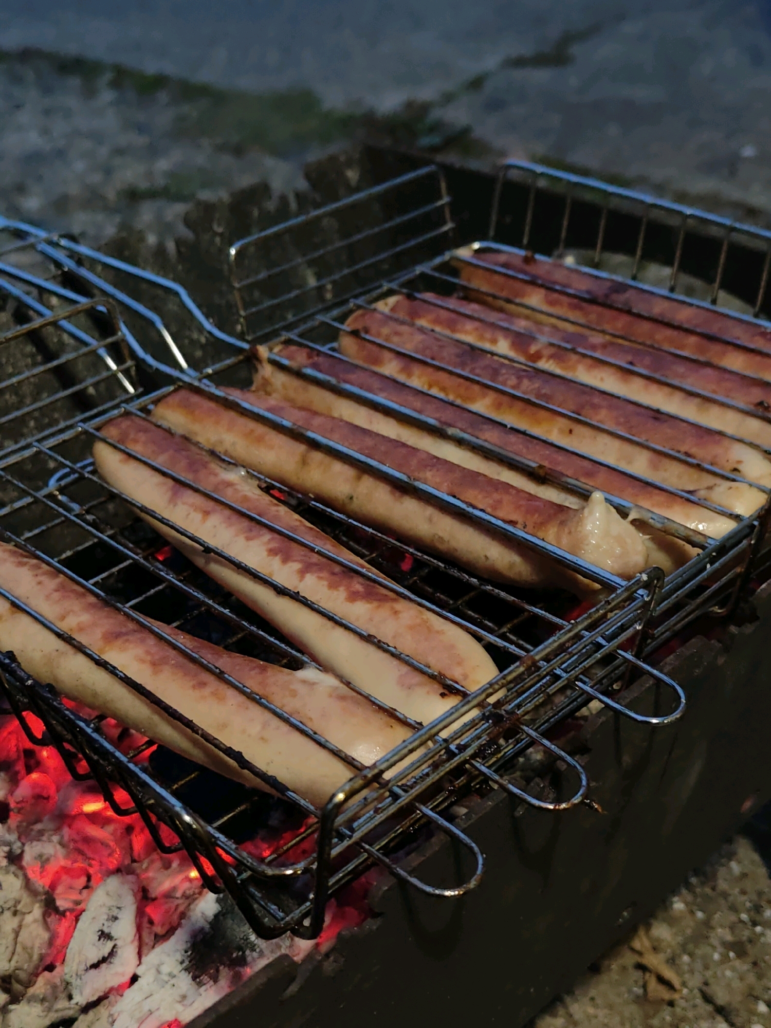 The image shows several sausages cooking on a grill over hot coals. The grill is a metal basket with a handle, designed to hold multiple sausages. The sausages are elongated and appear to be a light brown color, some with slight charring from the heat. Red-hot embers and gray ash are visible beneath the grill, indicating an active fire. The background is dark and out of focus, suggesting it is nighttime or a dimly lit outdoor area. The setting appears to be a backyard or campsite, with pebbles and dirt visible on the ground. No people or specific landmarks are identifiable in the image, making it impossible to ascertain the location within Chaikovsky, Russia.