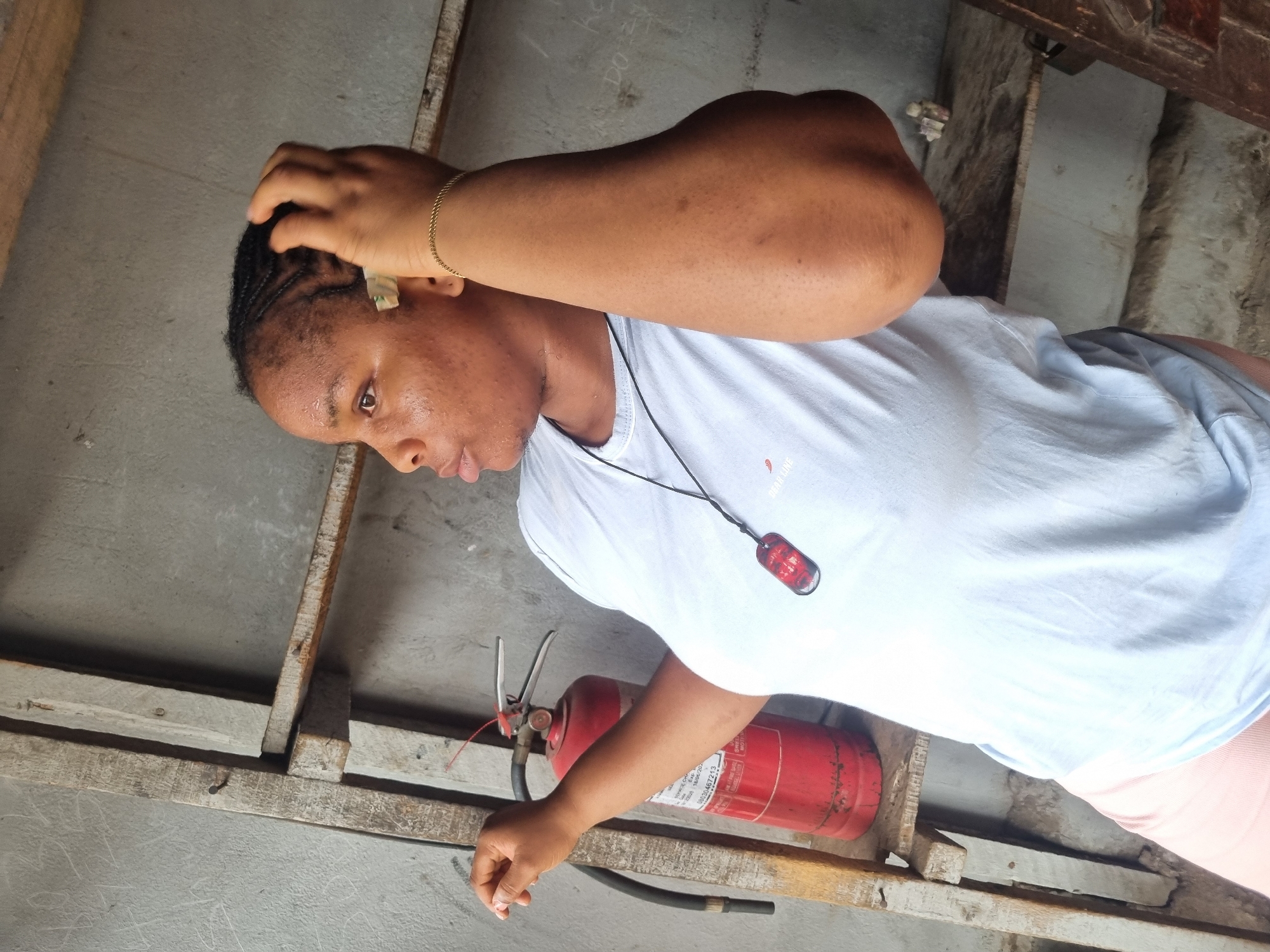 A medium shot shows a woman in Onitsha, Nigeria, wearing a white t-shirt and a gold bracelet, standing in what appears to be an unfinished or dilapidated structure with a grey concrete floor and a wooden frame. A red fire extinguisher rests on the wooden frame near her. She is touching her head with her hand and looking downwards. A black cord with a red ornament hangs around her neck.