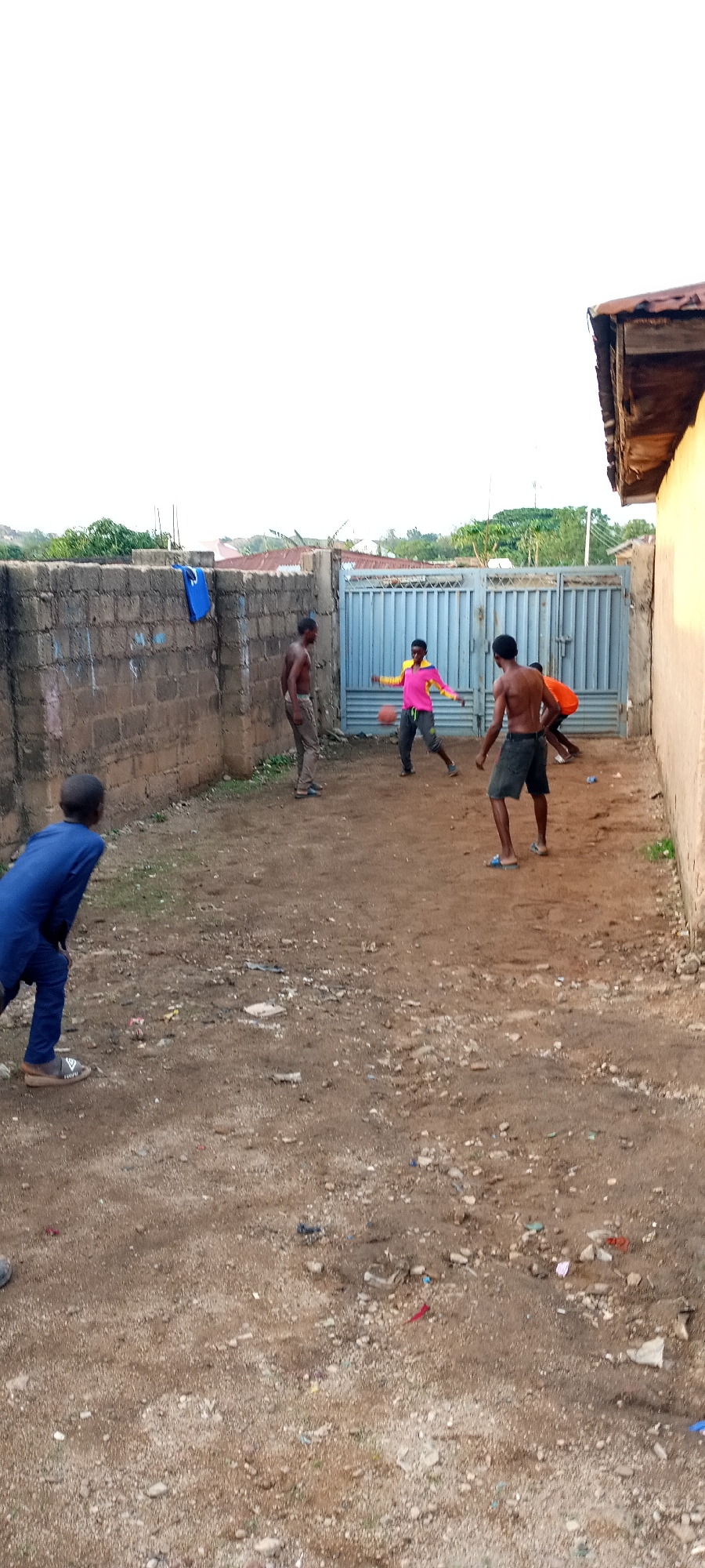 A group of approximately five young males, possibly adolescents or young adults, are engaged in an informal game, likely basketball or football, in an unpaved outdoor area. The central figure, wearing a vibrant pink and yellow long-sleeved shirt and grey pants, is dribbling a small orange ball. To his left, a shirtless individual in light brown pants stands watching, while another shirtless figure in dark shorts stands with his back to the viewer on the right. A person in an orange shirt is partially visible behind the latter. In the left foreground, a boy in a blue traditional outfit is bent forward, looking towards the game.
The scene is set in a dusty, dirt ground area, characteristic of an informal playing space in a residential environment. A sturdy concrete block wall borders the left side, with a blue shirt or jersey hanging on it. A large, grey metal gate forms the backdrop, beyond which trees, some distant rooftops, and scattered buildings suggest a semi-urban or residential area. On the right, a lighter-colored building wall with a worn, wooden roof structure is visible. The lighting indicates late afternoon or early evening, with soft, ambient light and a warm glow on the right wall. The overall atmosphere suggests casual play and community interaction in Jos, Nigeria. No visible text can be discerned in the image.