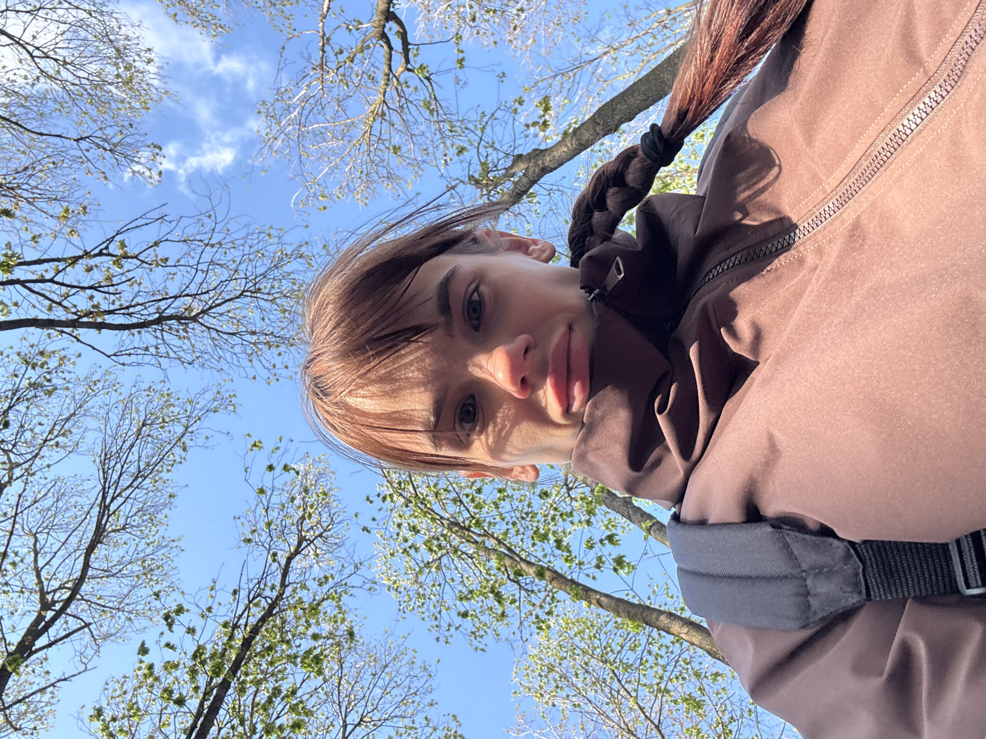 A low-angle, selfie-style shot captures a young person looking up, with a clear blue sky and trees as the background. The individual, who appears to be female, has light brown hair with bangs, and a long braid secured with a black hair tie visible on the right. She wears a brown jacket with a prominent zipper and a dark strap, possibly from a backpack, over her shoulder. Her lips are slightly puckered, suggesting a subtle expression.
The sky is bright and blue with a few scattered white clouds. Numerous tree branches frame the sky; some are bare, while others exhibit nascent light green leaves, indicating early spring. The bright sunlight suggests it is daytime, likely a sunny morning or afternoon. The setting appears to be an outdoor natural environment such as a park or forest, consistent with the early spring season in Rostov-on-Don, Russia. No visible text is present in the image.