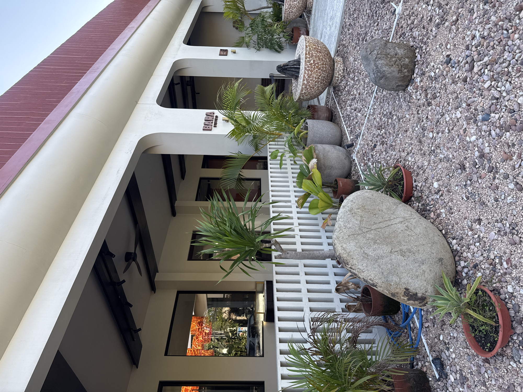 The image depicts the exterior of a building with a covered walkway, likely a restaurant or cafe, in North Kuta, Indonesia. The facade is cream-colored with arched openings into the walkway. A red, corrugated metal roof extends over the top. The walkway is supported by substantial white pillars. White slatted fencing runs along the front of the building, separating the walkway from an outdoor area.
This outdoor area is paved with small, multi-colored pebbles. Several potted plants are strategically placed around the area, including a tall dracaena plant, various palms, and a small agave in a terracotta pot. Large decorative stones, one very prominent and smooth, and another smaller, rougher one, are situated on the pebble ground.
In the background, through the slatted fence and reflected in the windows, are hints of interior lighting and possibly more greenery. A sign on the building reads "BARA BISTRO". In the distance, one can see larger potted plants, and a mosaic-patterned bowl with what appears to be a dark sculpture or decorative element at its center. The sky is clear and blue, suggesting fair weather. The overall impression is one of a well-maintained, modern outdoor dining or waiting area.