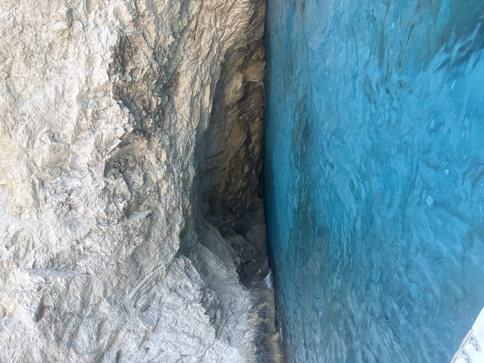 An overhead view captures a bright blue cave carved into a massive, textured white cliff face. The cave opening is dark and shadowed, contrasting with the luminous, clear blue water that fills the foreground. Ripples on the water's surface suggest gentle movement. The rock face is rough and layered, showing signs of erosion and mineral deposits. The scene appears to be a natural coastal formation.