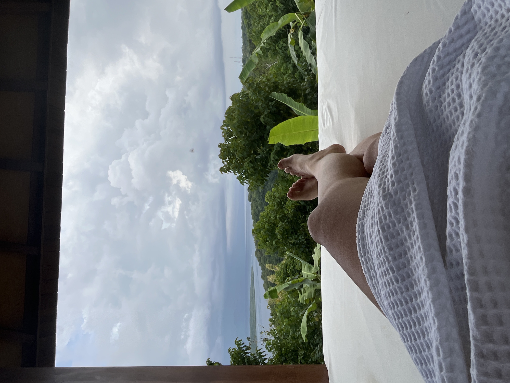 The image depicts a person reclining on a white surface, possibly a bed or lounge chair, with their legs crossed. They are partially covered by a textured white robe or blanket. The background reveals a view of lush green vegetation, including trees and banana plants. Beyond the greenery, there is a body of water and a cloudy sky. The scene is framed by the edge of a structure, possibly a building or deck, visible at the bottom and left side of the image. The location is likely Nusa Penida, Indonesia, due to the typical vegetation of banana trees and its hilly terrain facing the sea. The person is likely relaxing and enjoying the view.