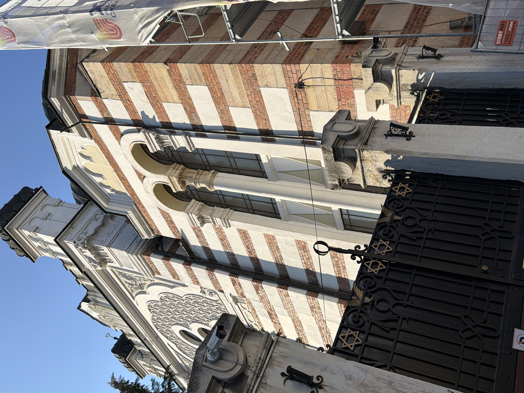 The image shows the exterior of a building, likely a synagogue, given the presence of the Star of David on the gate. The building's architecture features alternating stripes of brick and a light-colored material, possibly stucco. There are arched windows and ornate detailing on the facade. The sky is visible in the background. Scaffolding is covering part of the upper section of the building. A security camera is visible. A sign with "STROY RENT" is on the roof.