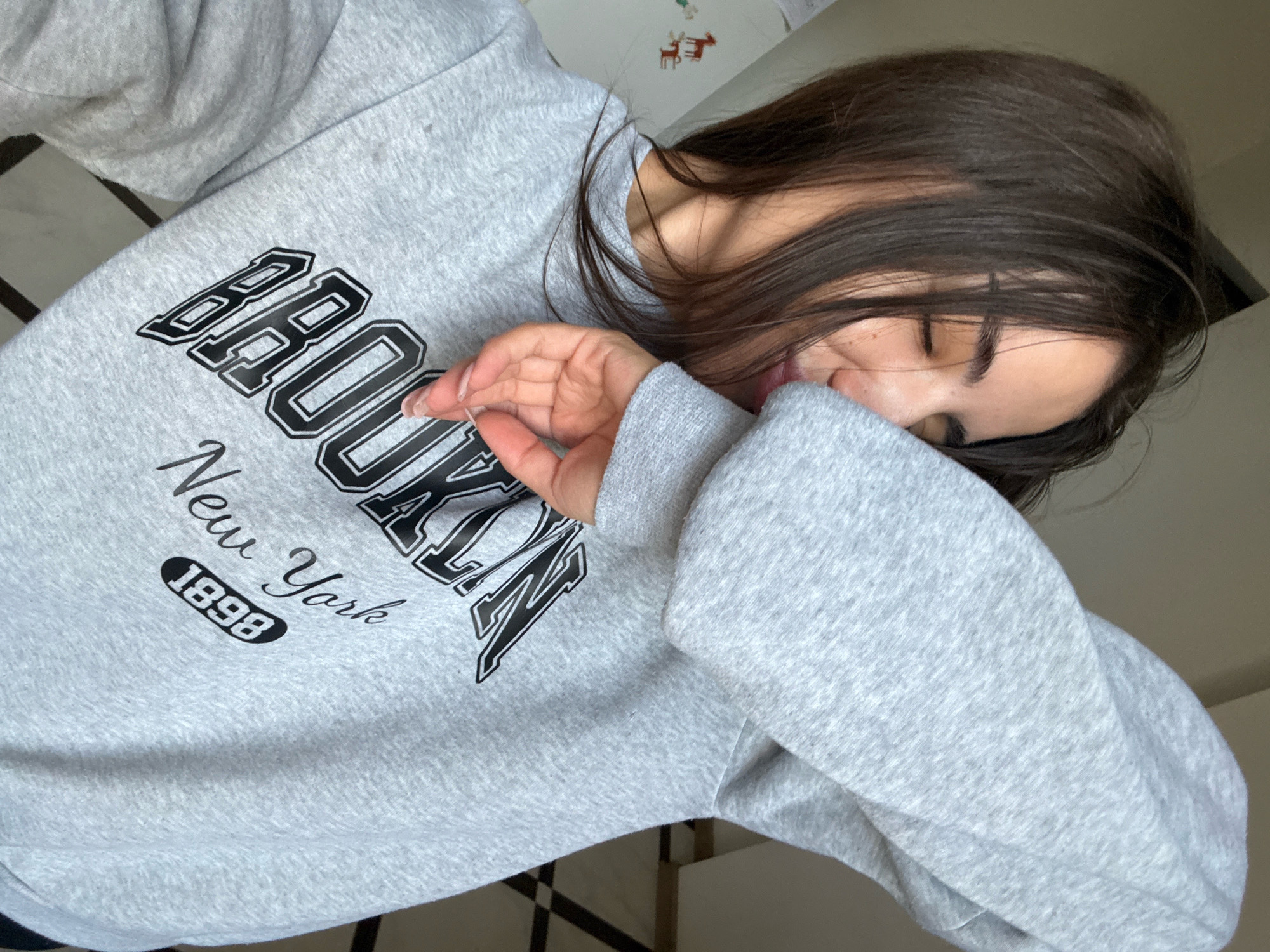 The image shows a young woman wearing a grey sweatshirt with "BROOKLYN New York 1898" printed on it. She is covering her face with her hand, obscuring most of her features. The background appears to be an indoor setting with a checkered floor. There are no identifiable landmarks or clues to suggest the location is in Chișinău, Moldova. The image is a close-up, focused on the woman and her sweatshirt, with limited environmental detail.