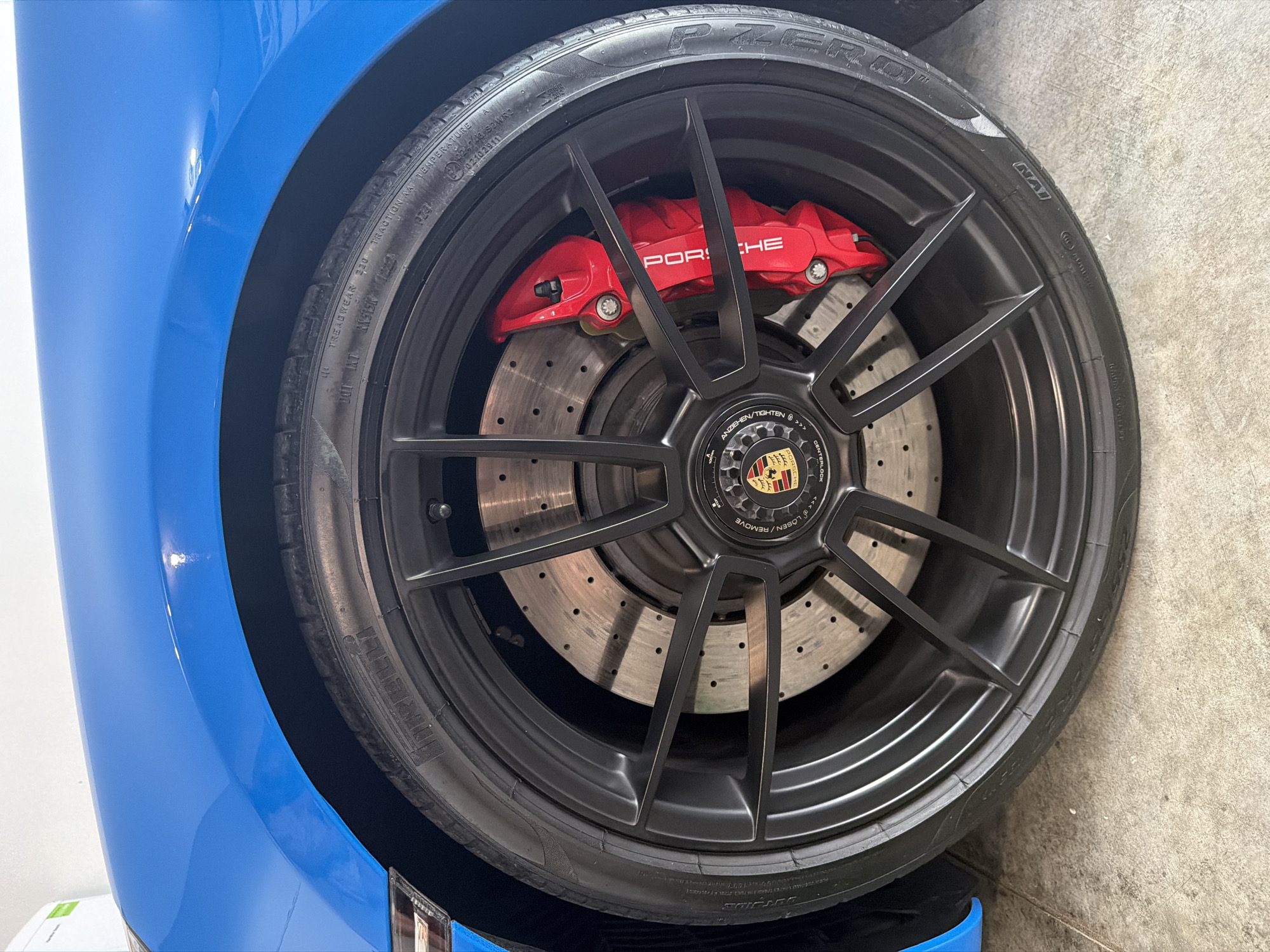 A close-up, low-angle shot shows a bright blue Porsche car's front wheel. The wheel is black with a multi-spoke design. A large red brake caliper with the word "PORSCHE" printed in white is visible. The brake rotor has numerous small holes. The tire is black and has text on its sidewall. The car is parked on a concrete surface. The background consists of a white wall and a section of the car's blue fender. The location is Wisconsin Dells, United States of America.