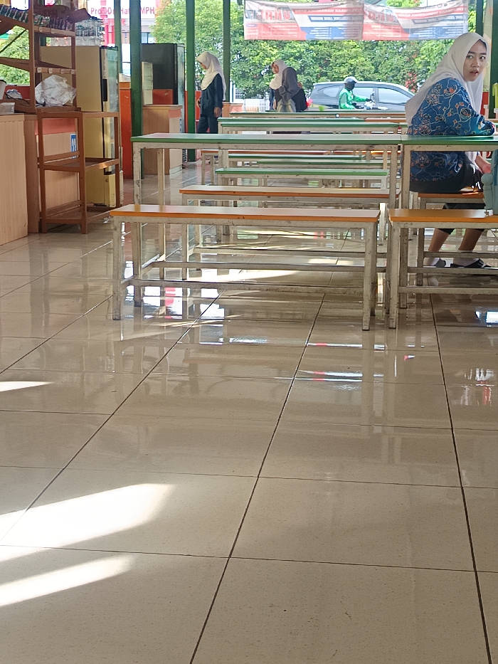 Inside a bright, tiled room, rows of empty tables and benches line the space. Two women wearing hijabs are present; one is seated at a table, her back turned, while the other walks further away. Outside, trees and a road with passing vehicles, including a motorbike, are visible through the open space. Sunlight streams onto the reflective floor, creating bright patches. A tall shelving unit stands on the left side of the room.