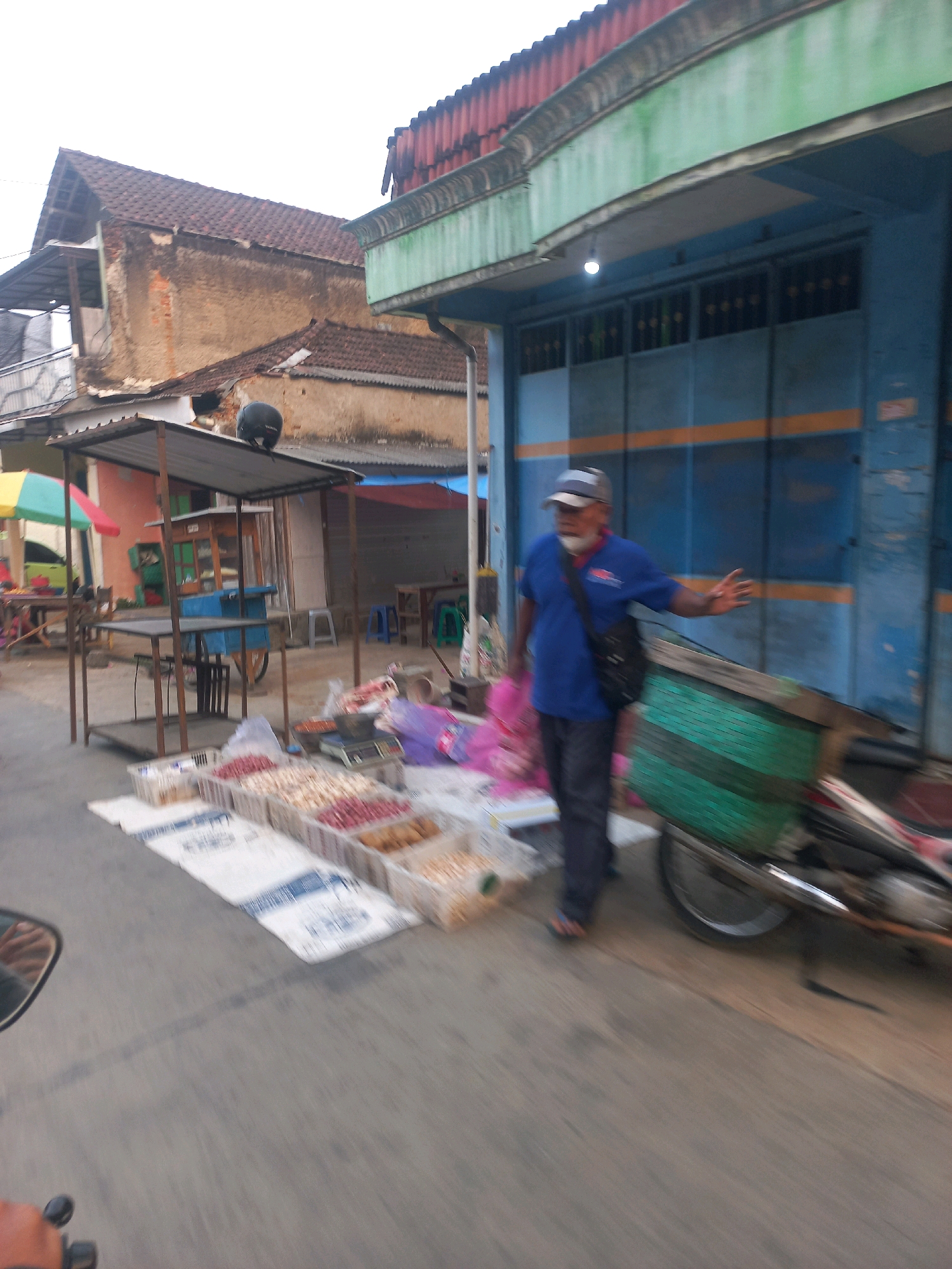 This is a motion-blurred photograph of a street scene in Sukoharjo, Indonesia. The foreground shows a motorcycle mirror and handlebar. The midground features a market stall with various goods, including what appear to be nuts and dried food items in plastic containers. A man wearing a blue shirt, a cap, and a sling bag is walking towards the right of the frame, with his right hand outstretched. Behind the stall, a blue building with orange stripes is visible, possibly a shop or warehouse. In the background, there are buildings with tiled roofs, suggesting a residential or commercial area. The overall impression is of a bustling street market.