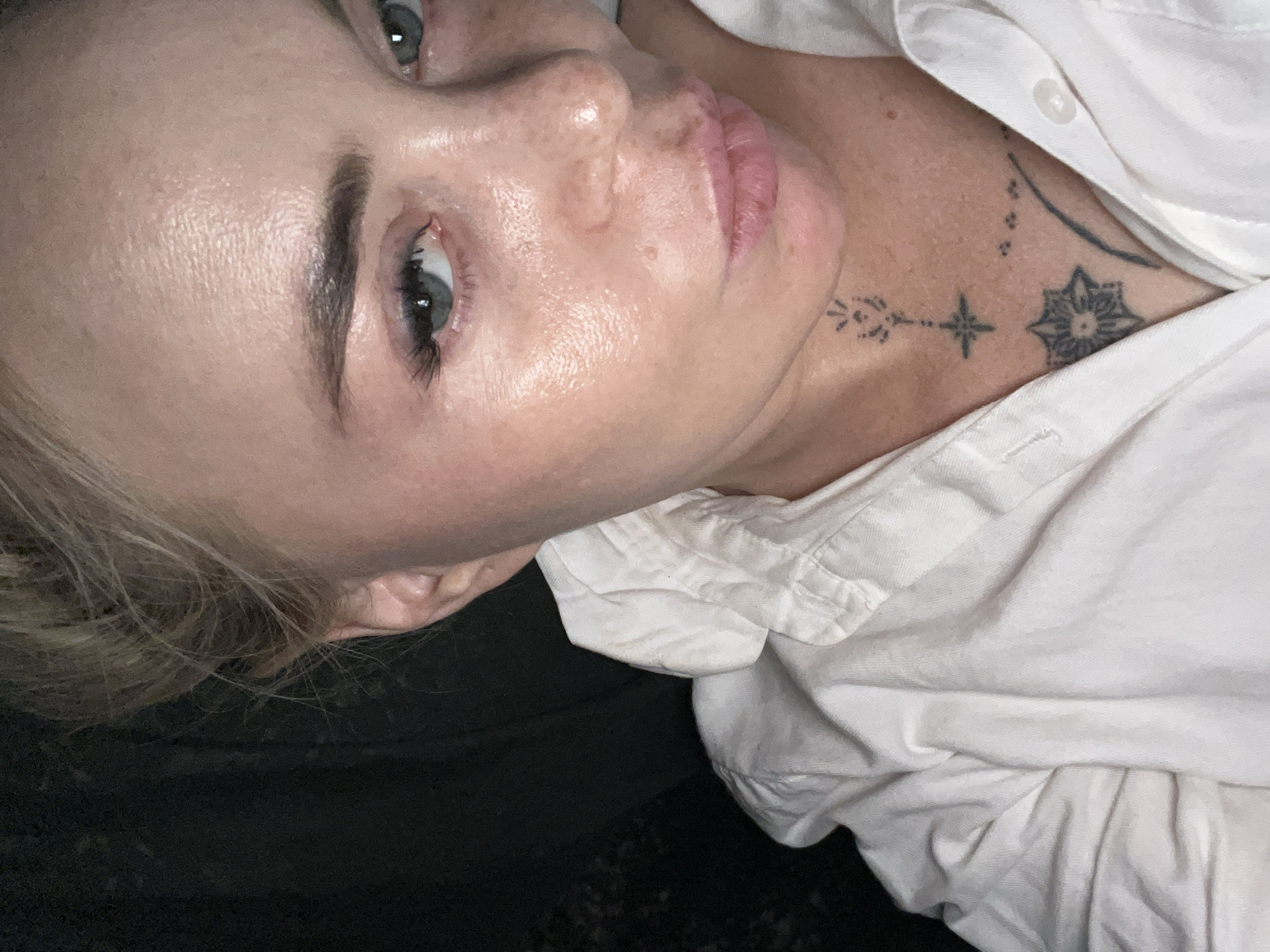 A medium shot shows a woman with blonde hair pulled back. She is wearing a white collared shirt. Her face is turned to her right, and her lips are pursed. Tattoos are visible on her neck and chest. The background is dark and out of focus.
