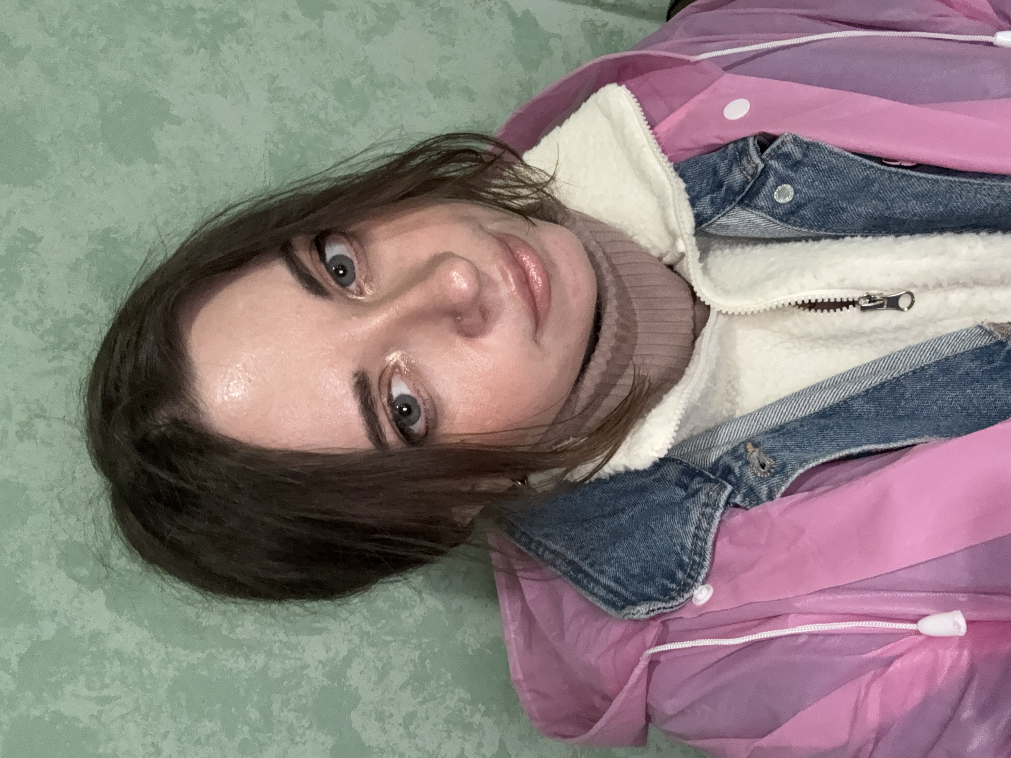 A woman with fair skin and dark brown hair is facing the camera. She has gray eyes, brown eyebrows, and a nose piercing. She is wearing a light brown turtleneck sweater, a white fleece-lined denim jacket, and a translucent pink raincoat. The background appears to be a green, slightly textured surface. The photograph does not contain sufficient information to determine a location in Moscow, Russia.