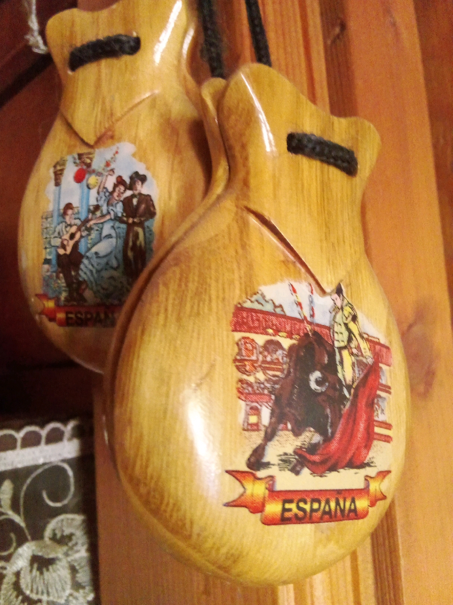 This is a close-up, eye-level, indoor shot of two wooden castanets, a traditional Spanish percussion instrument. The castanets are light tan, with a smooth, varnished surface. Each castanet is adorned with a colorful decal.
The castanet in the foreground features a decal depicting a bullfight. A matador in a yellow and green suit stands to the right, holding a red cape. A dark brown bull is positioned in the center, its head lowered. The background of the decal shows a bullring with red and yellow striped railings. A banner across the bottom of this decal reads "ESPANA" in red letters on a yellow and red striped background.
The castanet in the background has a decal showing a group of people in traditional Spanish attire, perhaps dancing or celebrating. One figure is playing a guitar. This decal also has text at the bottom, though it is partially obscured.
Both castanets are suspended by black cords. The background consists of vertical wooden planks, likely part of a wall or furniture. The lighting is warm and slightly diffused, suggesting an indoor setting.
The image does not contain information about the city of Santomenna, Italy.