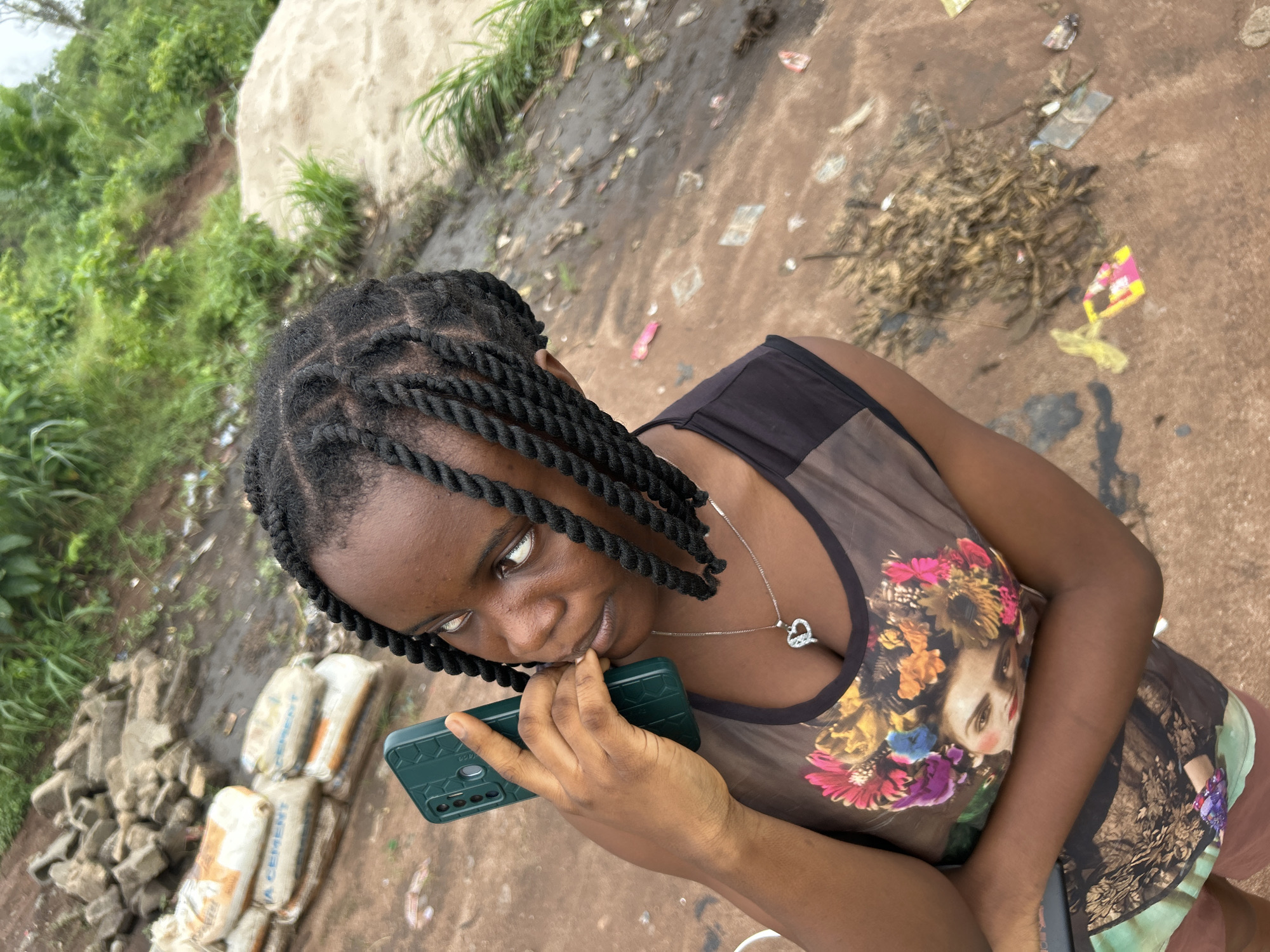 In Ihiagwa, Nigeria, a young woman with braided hair holds a green smartphone to her ear, her finger resting on her chin. She wears a dark, sleeveless top with a colorful graphic print of a woman's face adorned with flowers. A silver heart-shaped pendant hangs from a delicate chain around her neck. The background reveals an outdoor setting with scattered building materials, including bags of cement, piles of rocks, and a sandy mound. Patches of green vegetation and debris litter the ground, suggesting a construction or undeveloped area.