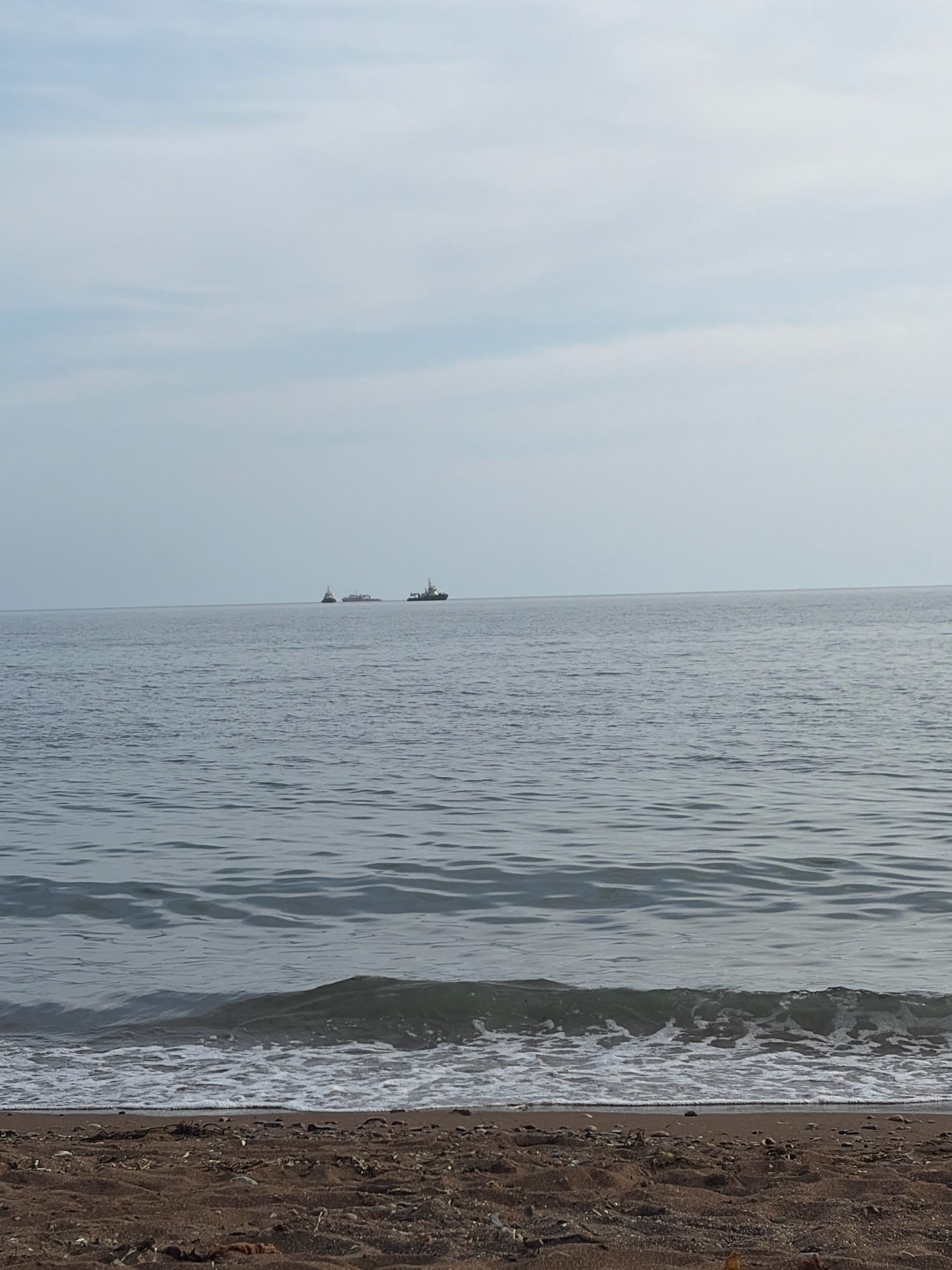 A wide shot captures a tranquil scene of the sea and beach in Korsakov, Russia, under an overcast sky. The foreground is dominated by a dark, sandy beach littered with small stones and debris. Gentle waves lap the shore, with white foam cresting as they break. The sea stretches out, a vast expanse of calm, grey water reflecting the muted tones of the sky. In the distance, three vessels are visible on the horizon. The closest to the viewer is a tugboat. Further out, a larger ship appears to be under tow by another vessel, though the details are indistinct. The overall impression is one of peaceful solitude.