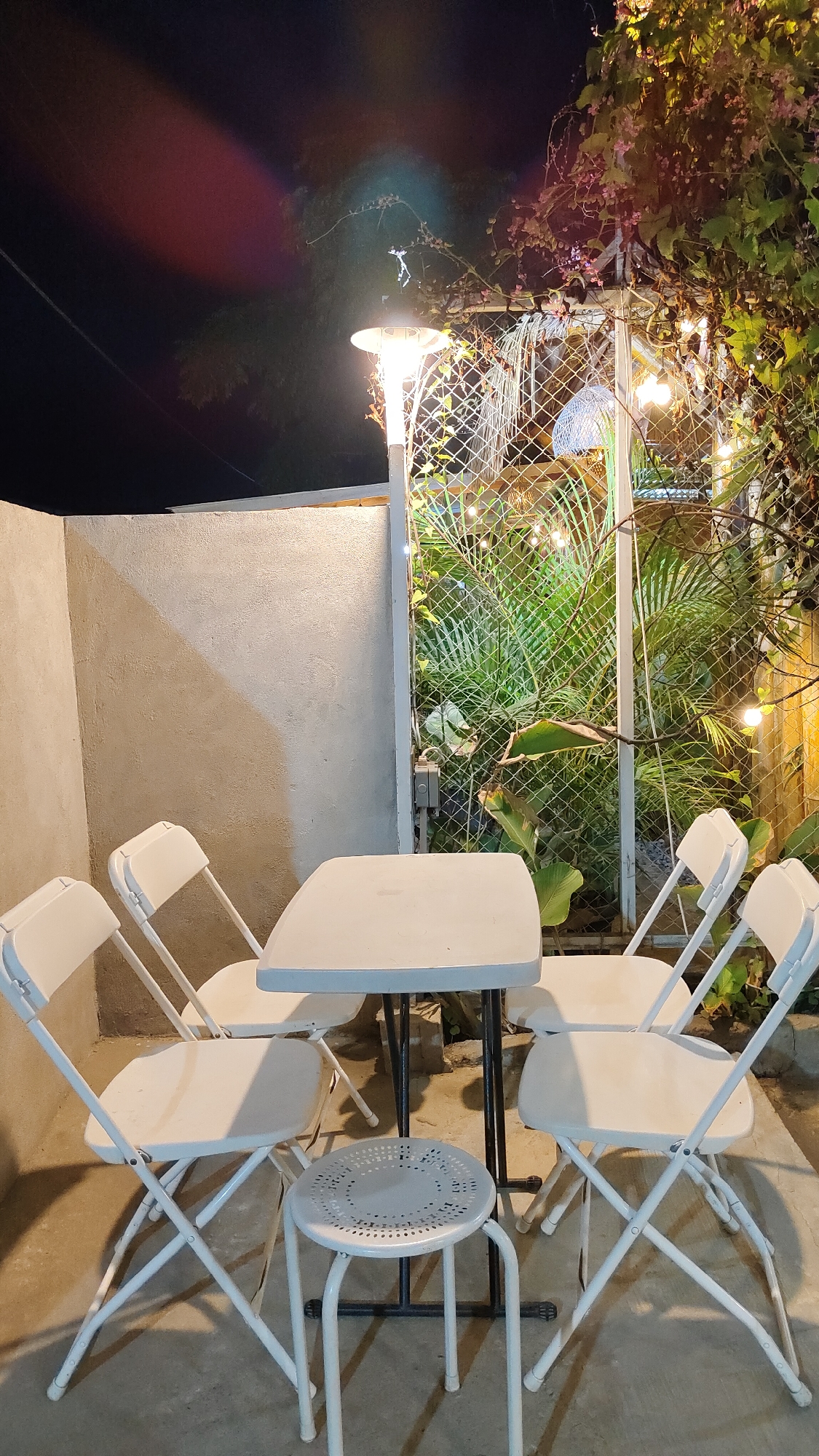 The image depicts an outdoor seating area at night, likely a cafe or restaurant, located in Keudebayu, Indonesia. A white rectangular table is centrally positioned, surrounded by four white folding chairs and a small white stool. Behind the table, a metal fence encloses a lush green garden with tropical plants. String lights are visible hanging above the garden, and a tall lamp post illuminates the scene. The ground is paved with concrete. The background shows a textured concrete wall and the dark night sky, with subtle light streaks suggesting artificial lighting effects. No people are present in the scene.