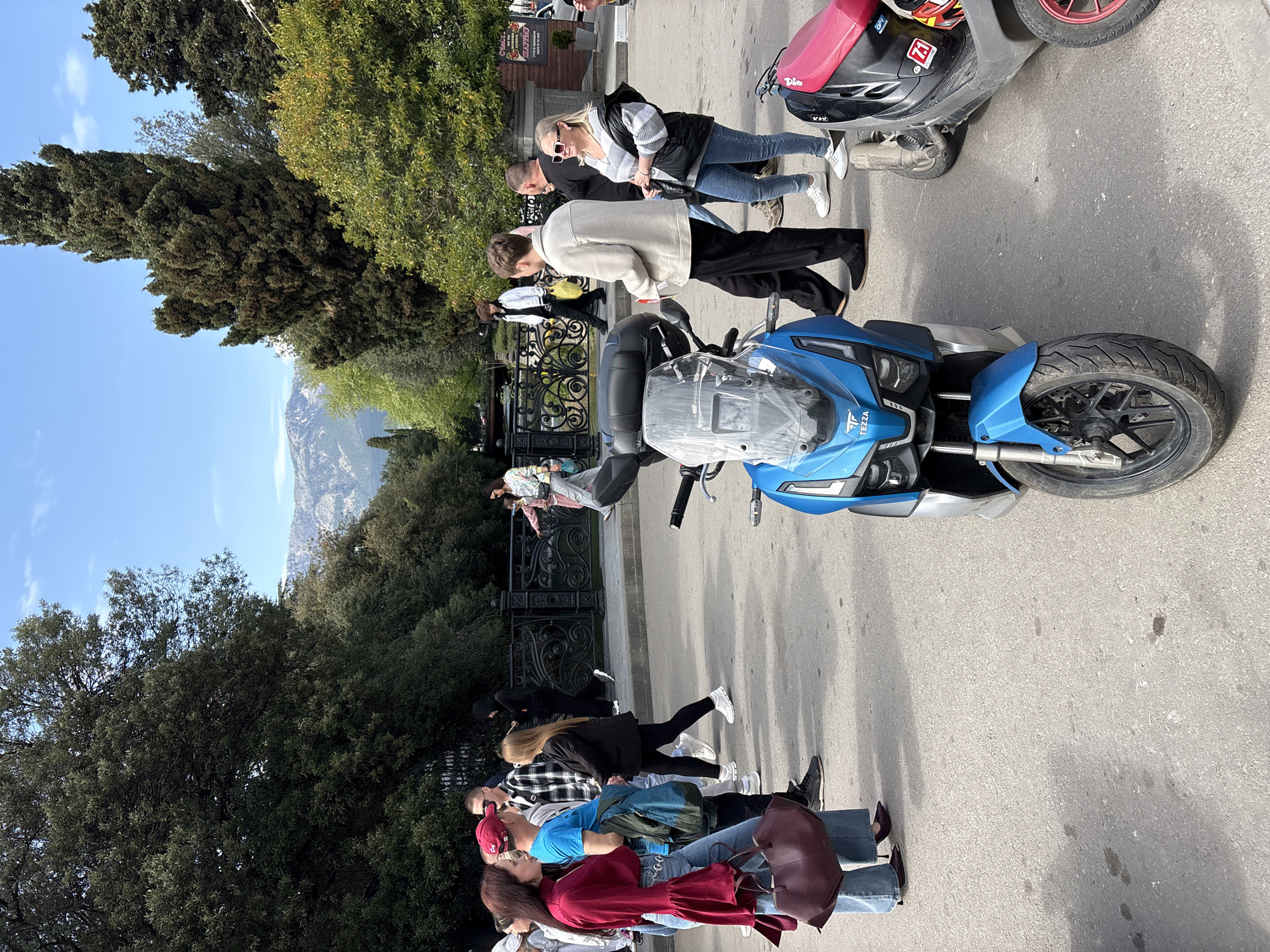 The image depicts a street scene in город Ялта, Ukraine, featuring a group of pedestrians and two parked motorcycles. The first motorcycle is a blue Tezza brand vehicle and the second is red and black in color. Pedestrians are walking on the sidewalk, near the decorative metal fence of a park or garden. The location is urban, featuring trees, shrubs, and a distant mountain. Weather appears to be sunny.