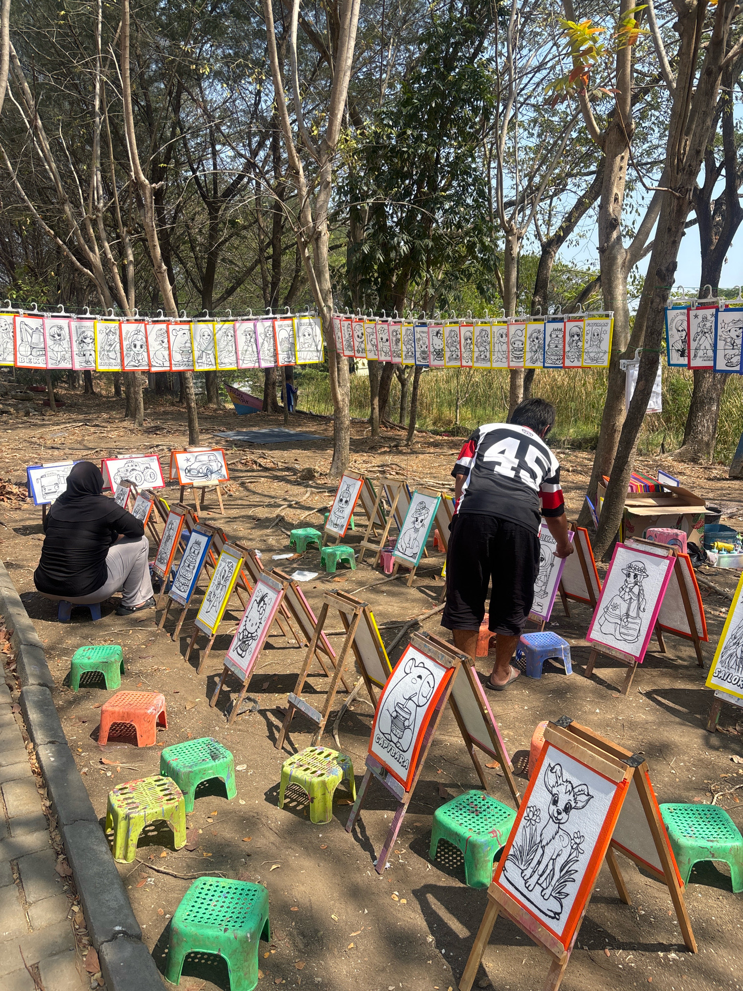 This image depicts an outdoor art activity area, likely in Tegal, Indonesia, under a canopy of trees. A row of coloring pages featuring cartoon characters and animals are hung from a wire, suspended between trees. In the foreground, several children's easels are set up, each displaying a black and white drawing on a white background with colorful borders. Some of these drawings are identifiable as Hello Kitty and a deer. Small plastic stools in green and orange are scattered around the easels. Two individuals are present: one person, wearing a black hijab and grey pants, is seated and facing away from the camera, looking at a drawing on an easel. Another person, wearing a black and white jersey with the number 45 and black shorts, is standing and appears to be interacting with the easels. The ground is covered in dirt and fallen leaves.