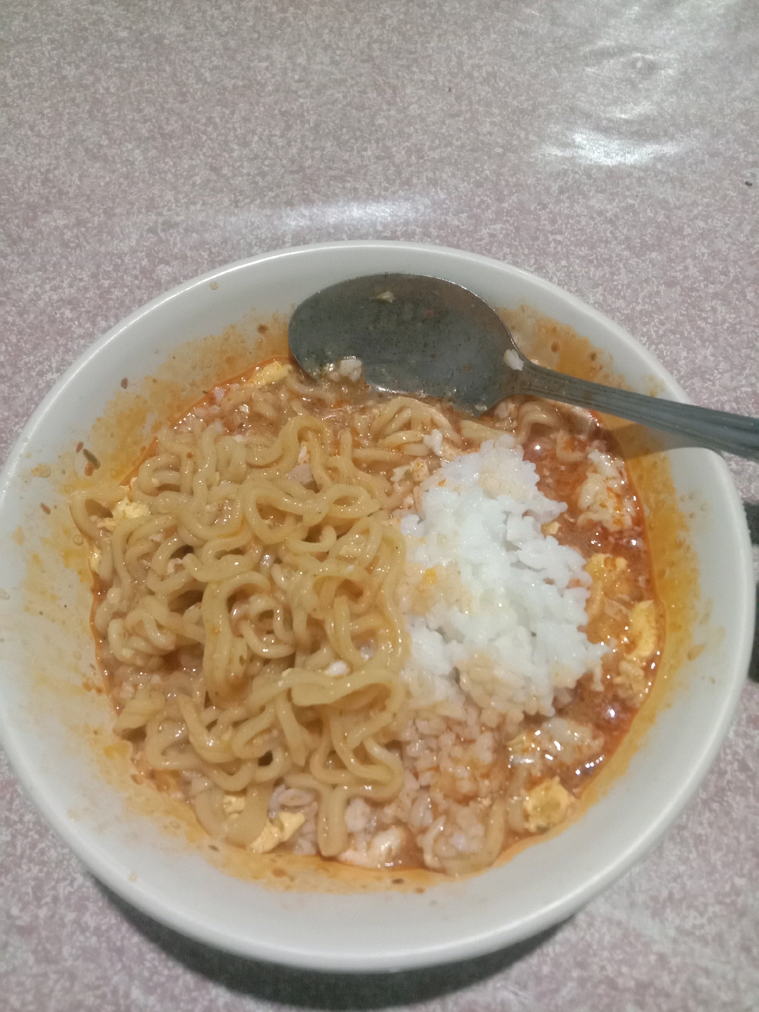 A close-up, high-angle shot shows a white bowl of ramen noodles mixed with rice and a reddish-orange broth. A silver spoon is resting on the rim of the bowl, its handle extending out of frame to the right. The ramen noodles are tightly curled and appear to be coated in the sauce. A pile of white rice sits on the right side of the bowl, partially obscuring the noodles. The broth has a slightly oily sheen and appears to contain small pieces of egg. The bowl is placed on a patterned tablecloth with a light beige and white speckled design. The lighting is dim and appears to be artificial, casting soft shadows. The background is out of focus, showing more of the patterned tablecloth.