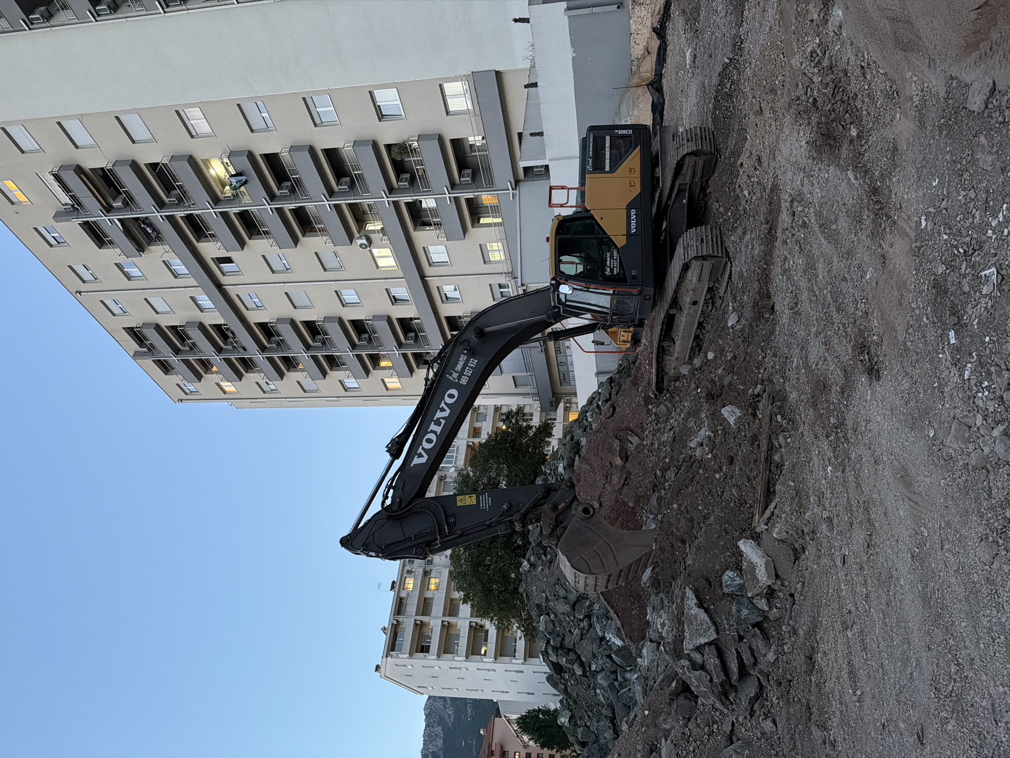 The image depicts a Volvo excavator parked on a construction site, set against the backdrop of an urban landscape. The excavator, primarily black and yellow, is positioned on a pile of dirt and rocks. In the background, multi-story residential buildings rise, with some windows illuminated, suggesting either dusk or early evening. The sky is a clear blue, transitioning to a lighter hue near the horizon. The construction site appears to be a new development or renovation area. The buildings and excavator indicate an urban setting, possibly the city of Budva, Montenegro. The presence of construction equipment suggests active work in progress.
