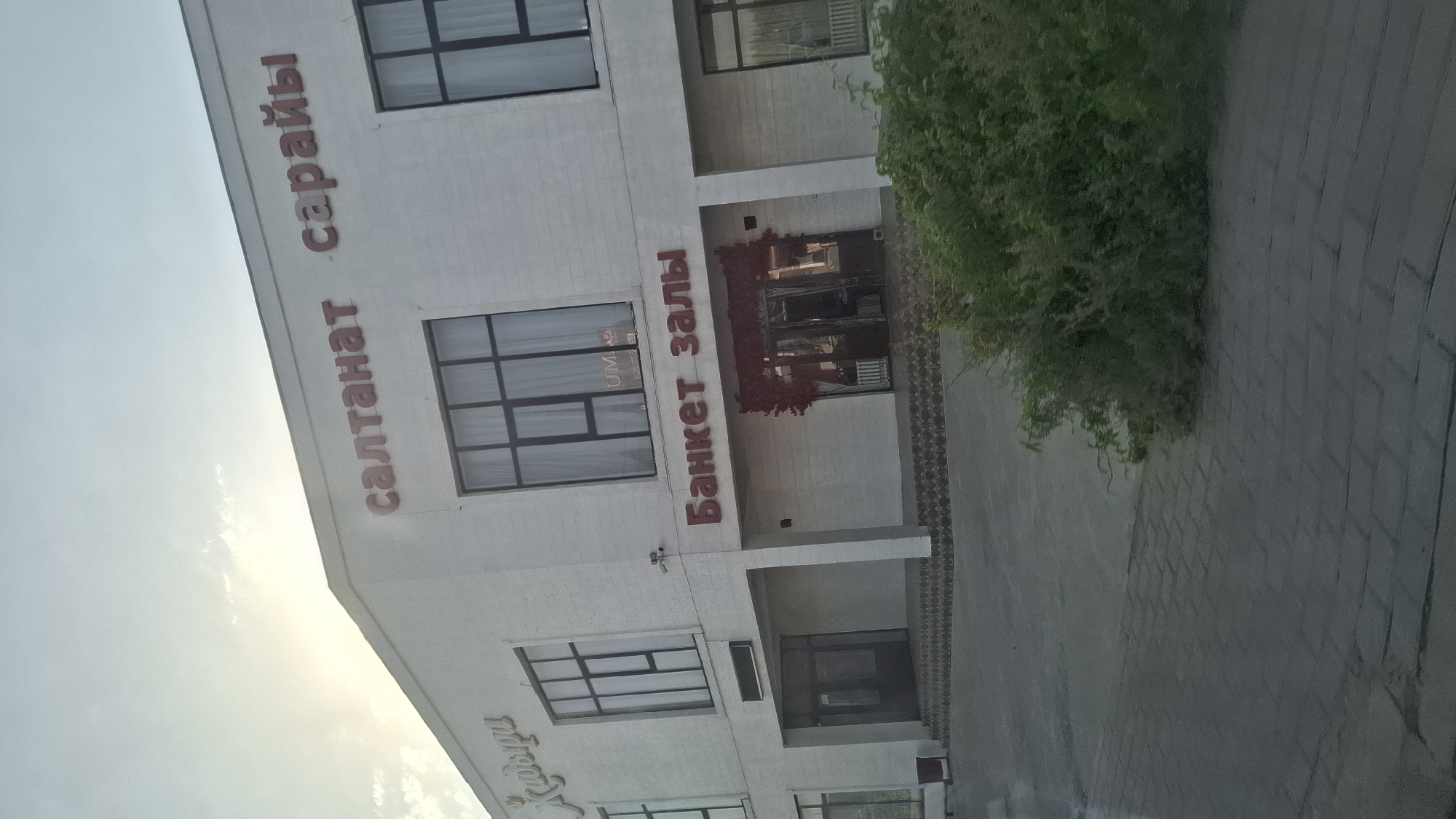 The image shows the exterior of a modern building with the name "САЛТАНАТ САРАЙЫ" in large red letters on its facade. Below this, also in red letters, is "Банкет залы", indicating it is a banquet hall. The building appears to be a single or two-story structure with large windows. There are entrances to the building, one of which is decorated with red floral arrangements. To the left of the main entrance, there's a sign that is partially visible and appears to be in Cyrillic script. The building is situated in a location that suggests it is in a city, with paving stones visible in the foreground and a patch of green bushes to the right. The sky is visible above the building, with some clouds and the sun peeking through. The overall impression is that of a commercial establishment, likely a venue for events, located in Шұбарсу, Kazakhstan.