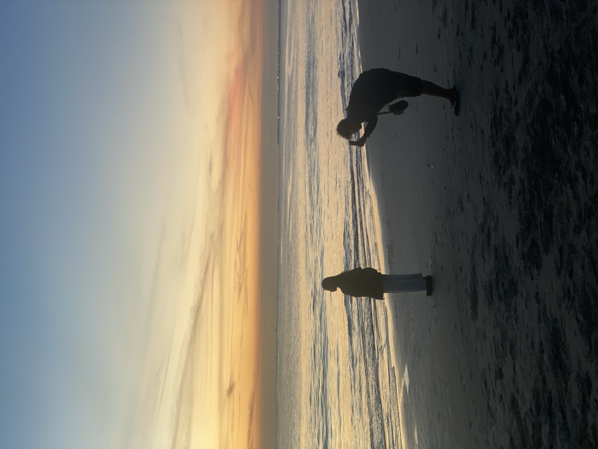 Two people are on a beach during sunset. One is bending over with what appears to be a net. The other is standing and looking out at the water. The location is Kuta Selatan, Indonesia.