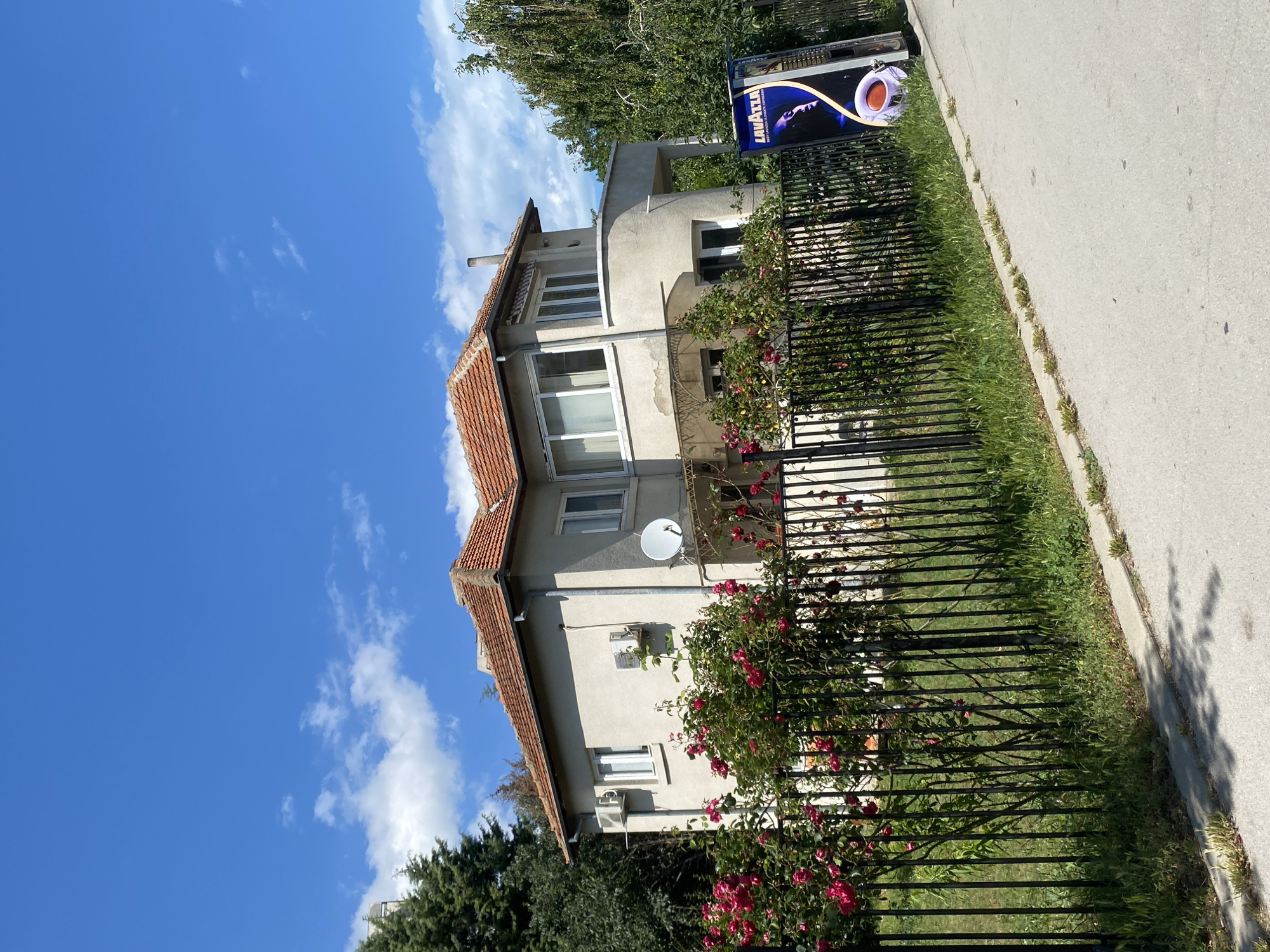 A two-story, light-colored house with a red-tiled roof is situated in the city of Varna, Bulgaria. The house features multiple windows, air conditioning units mounted on the exterior walls, and a satellite dish. A black metal fence encloses the property, behind which lush green grass and vibrant pink rose bushes are visible. To the right of the house, a tall, slender advertisement poster for Lavazza coffee stands next to the fence. The sky is a clear blue with scattered white clouds, and a paved road runs along the foreground, casting shadows from unseen foliage. Trees surround the house, with a large, leafy tree visible to the right.